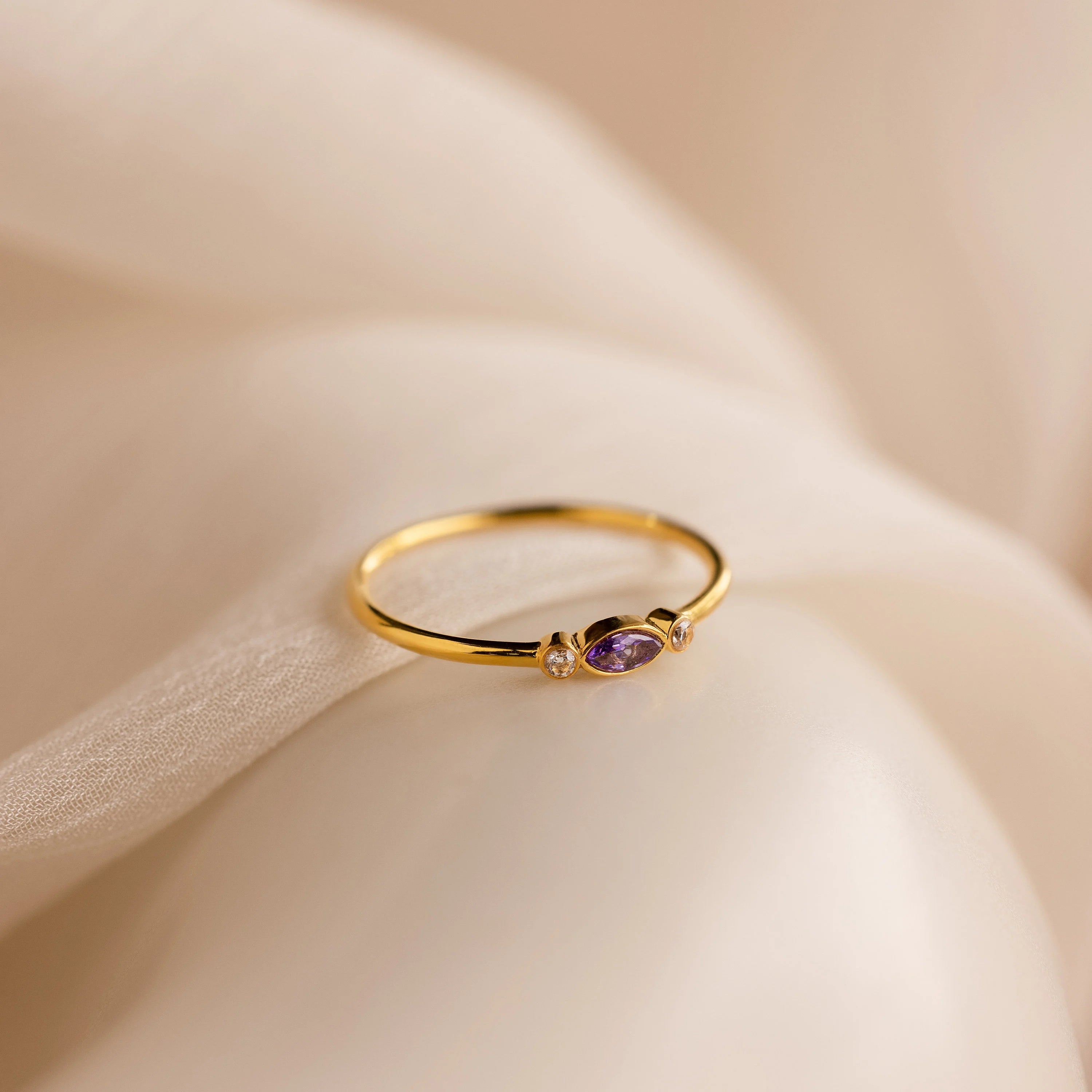 The Marquise Bezel Birthstone Ring showcases a marquise-cut birthstone, two sparkling diamonds, and a rich purple gemstone, elegantly displayed on soft cream fabric for a vintage-inspired look.
