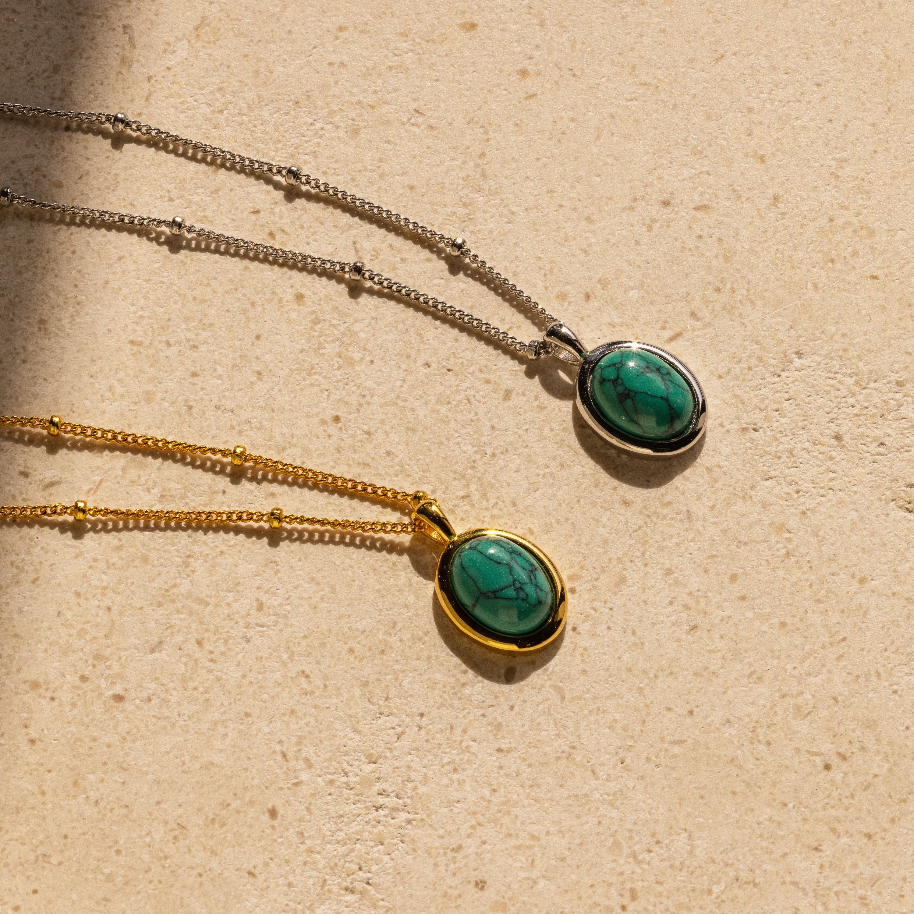 Two Turquoise Pendant Necklaces—one with a gold chain, the other with a silver chain—rest on a beige surface, offering a boho-chic touch to any outfit.