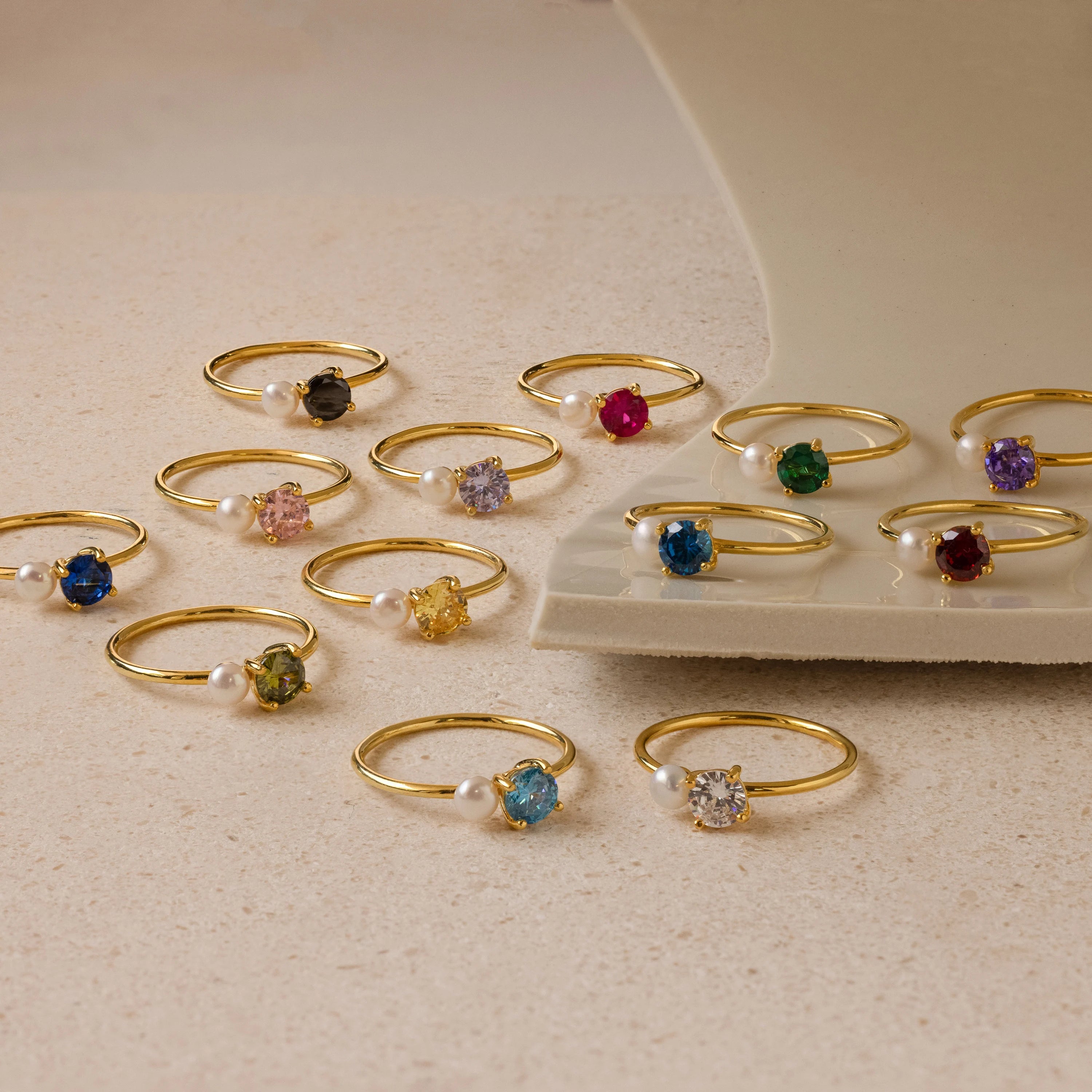 Birthstone & Pearl Rings, featuring elegant pearls and colorful gemstones, are displayed on a beige surface and a white ceramic plate.