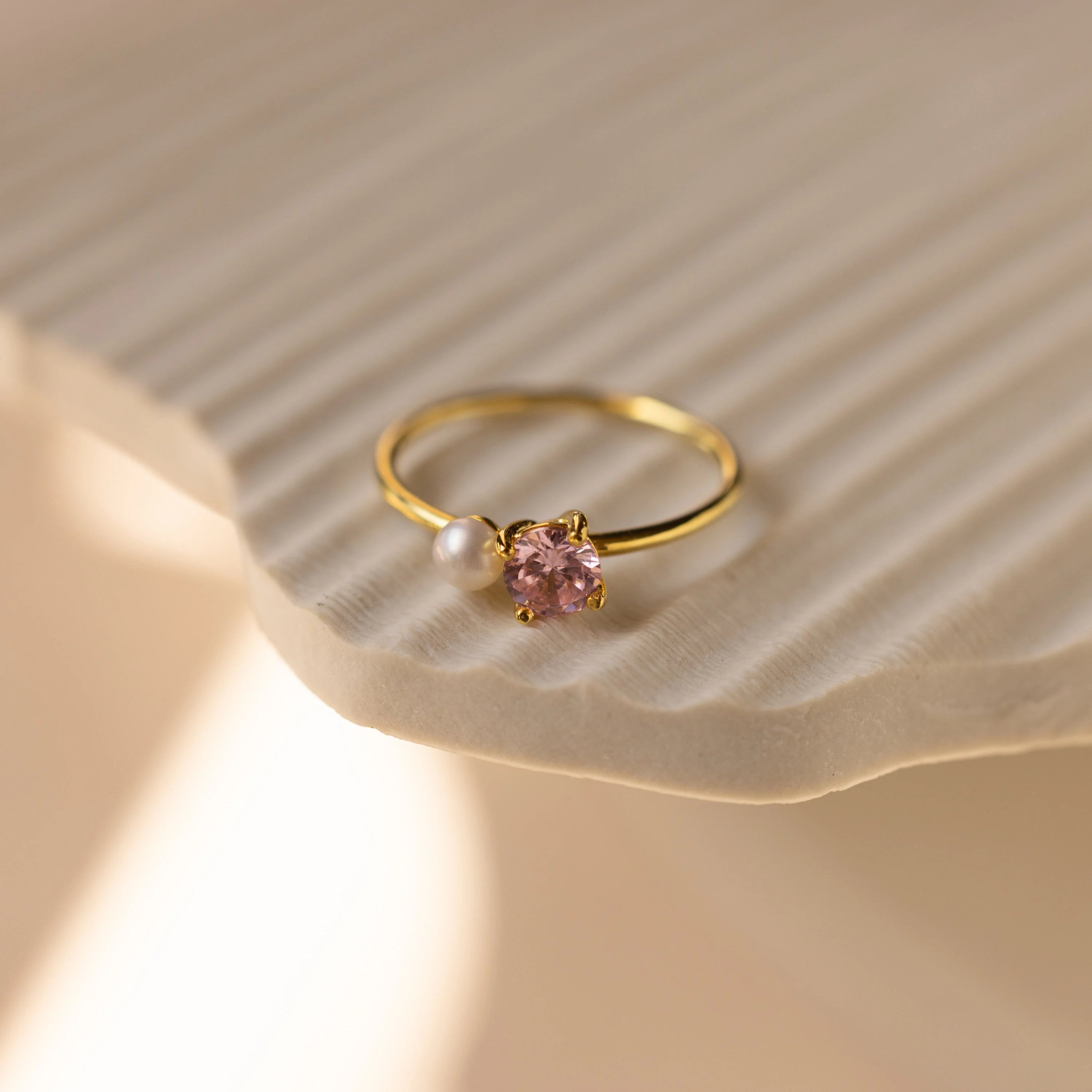 The Birthstone & Pearl Ring, featuring a pink gemstone and a delicate pearl set in gold, rests on a textured light surface—an elegant piece of personalized jewelry.