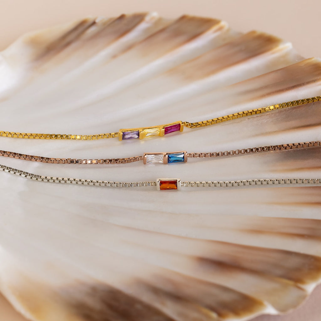 The Multiple Baguette Birthstone Bracelet features three delicate box chain bracelets with colorful rectangular gemstones, beautifully displayed on a seashell—ideal for stacking or creating your own custom birthstone look.
