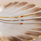 The Multiple Baguette Birthstone Bracelet features three delicate box chain bracelets with colorful rectangular gemstones, beautifully displayed on a seashell—ideal for stacking or creating your own custom birthstone look.