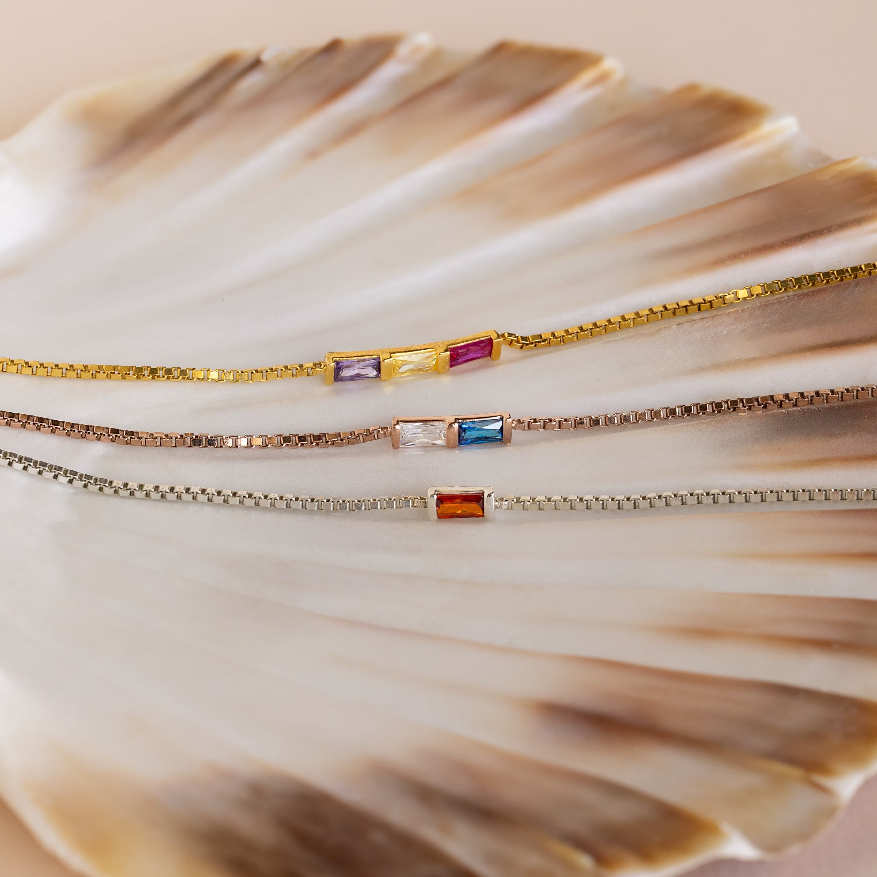 The Multiple Baguette Birthstone Bracelet features three delicate box chain bracelets with colorful rectangular gemstones, beautifully displayed on a seashell—ideal for stacking or creating your own custom birthstone look.