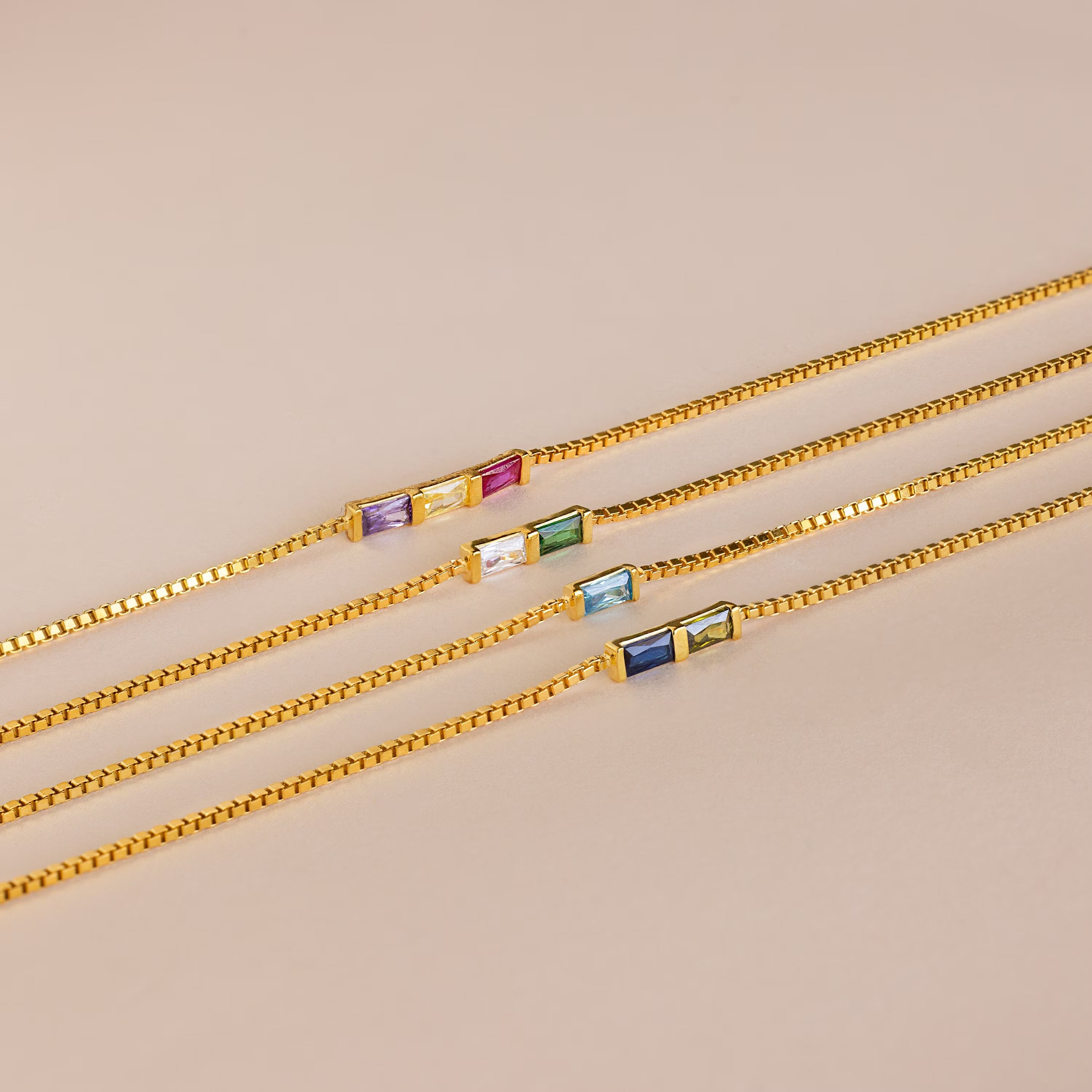 Four Multiple Baguette Birthstone Bracelets with gold box chains and two colorful rectangular gemstone charms each are displayed on a beige background—perfect as a custom birthstone bracelet gift.