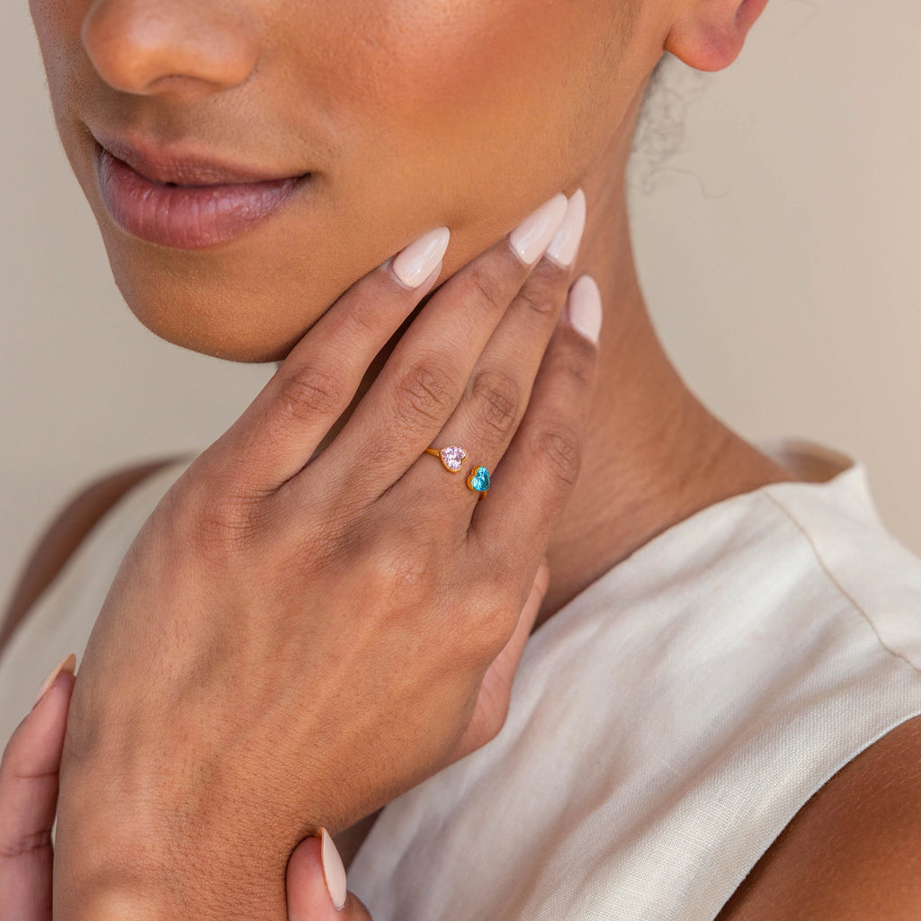 A woman in a sleeveless top displays her hand adorned with the Duo Heart Birthstone Ring—two heart-shaped gemstones set on a gold band, offering a unique and personalized touch.