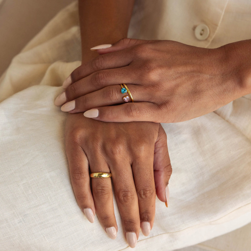 Two hands with manicured nails rest on cream fabric, adorned with gold rings featuring heart-shaped gemstones and a Duo Heart Birthstone Ring personalized with a custom charm.