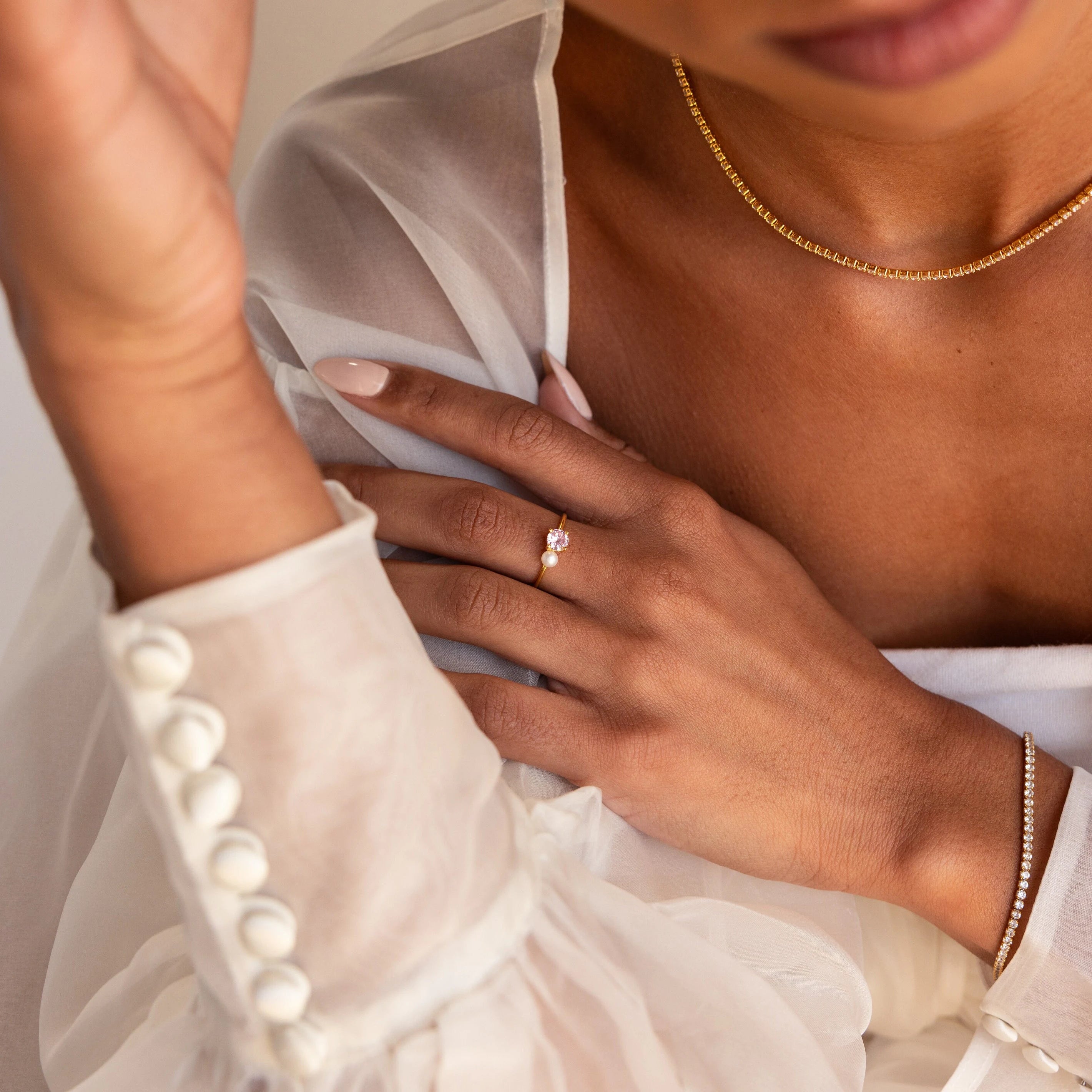 Woman wearing a sheer white blouse, gold necklace, bracelet, and the Birthstone & Pearl Ring, with light pink polished nails.