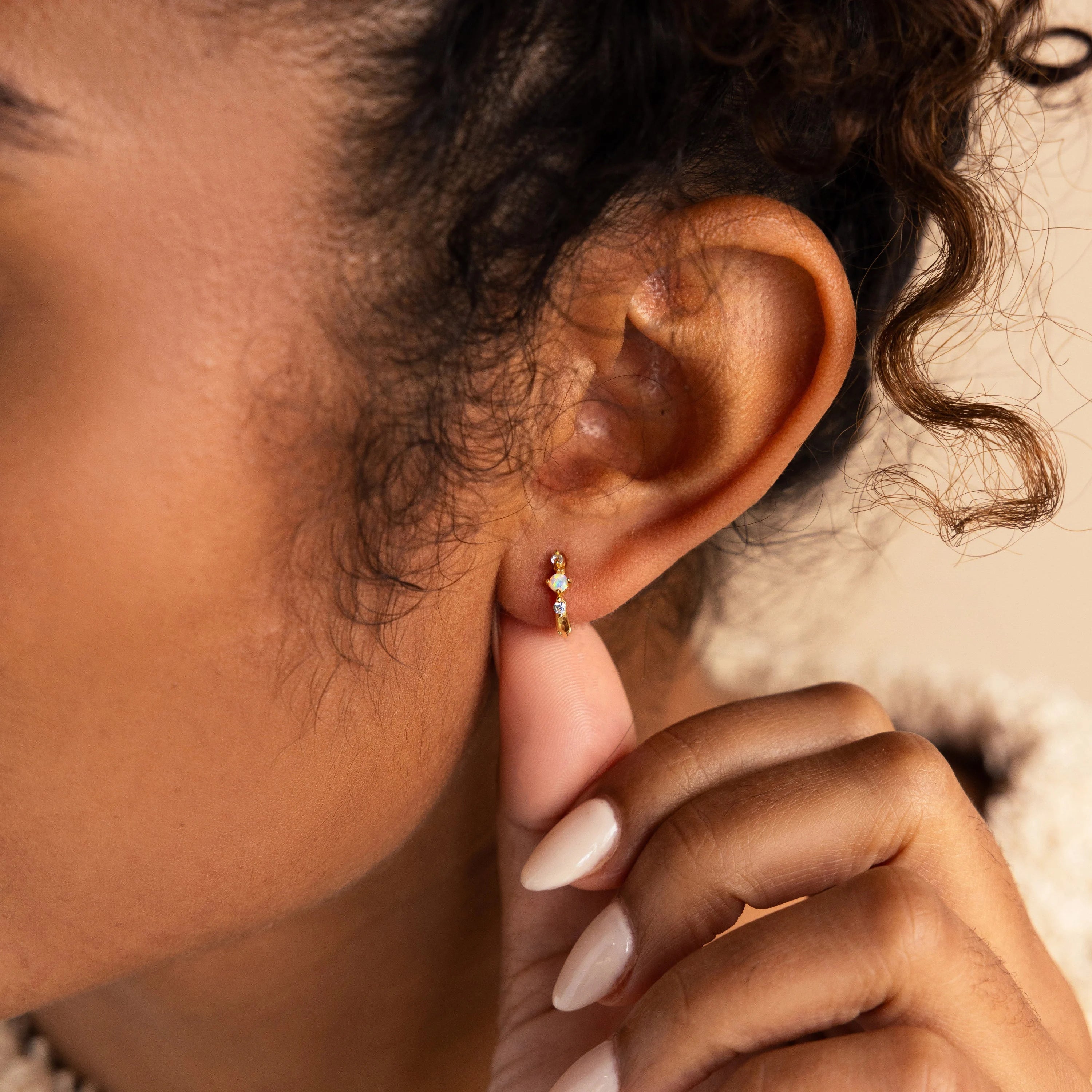 Close-up of a woman's ear adorned with a small gold huggie earring featuring three gemstones—an opal center stone flanked by two tiny diamonds—highlighting the earring's delicate and elegant design.