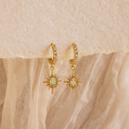 The Pave Opal Star Huggies capture celestial charm with gem-studded starburst pendants, beautifully showcased on a textured fabric background.