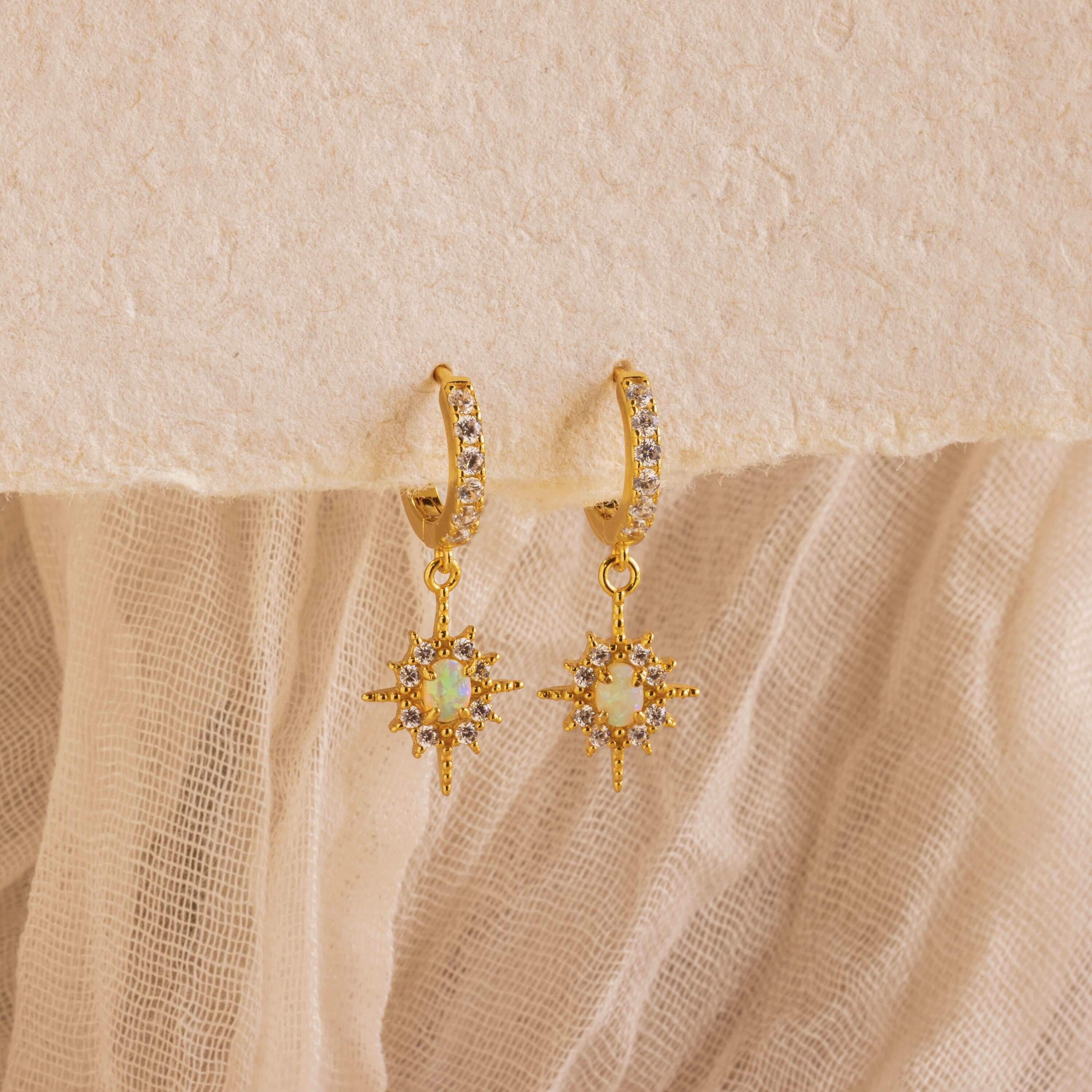 The Pave Opal Star Huggies capture celestial charm with gem-studded starburst pendants, beautifully showcased on a textured fabric background.
