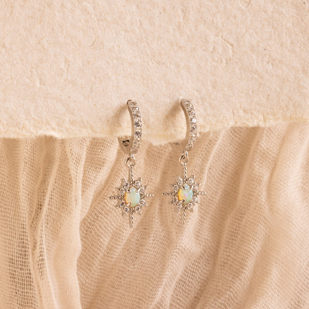 Pave Opal Star Huggies, sparkling silver hoop earrings with opal star pendants, are showcased on a textured beige fabric background.