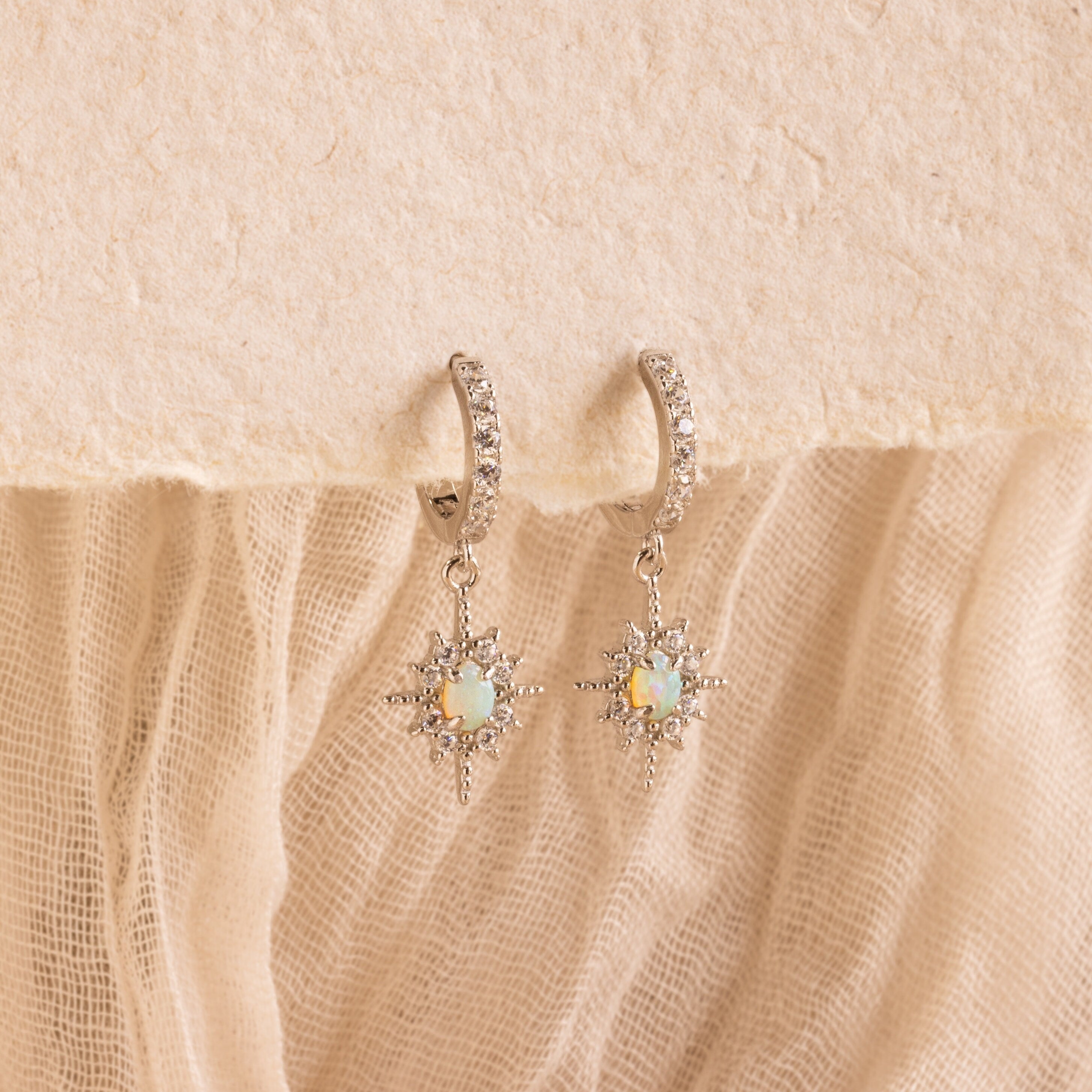 Pave Opal Star Huggies, sparkling silver hoop earrings with opal star pendants, are showcased on a textured beige fabric background.
