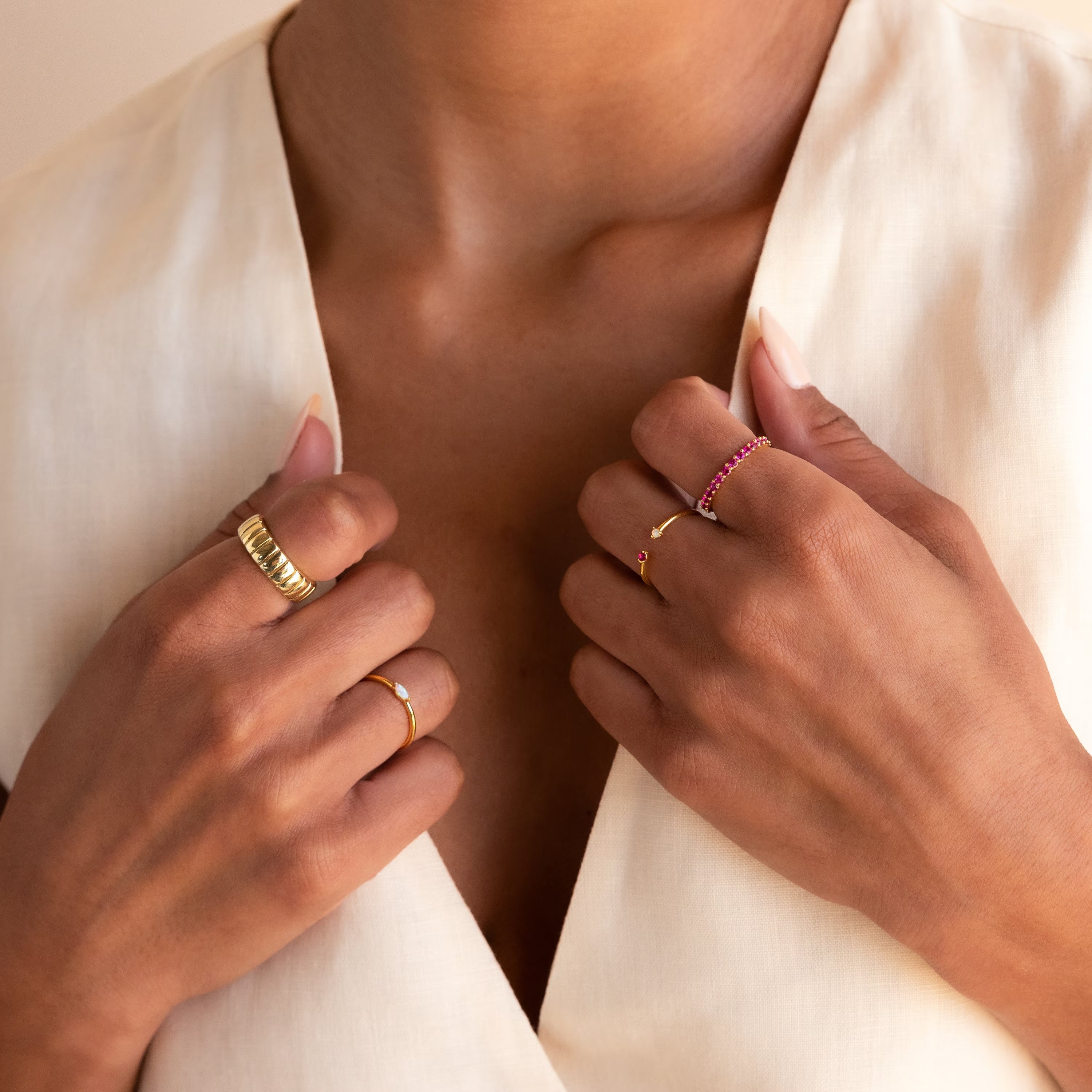 A woman in a cream blouse with a deep neckline showcases the Duo Birthstone & Opal Ring along with other gold and gemstone rings on both hands.