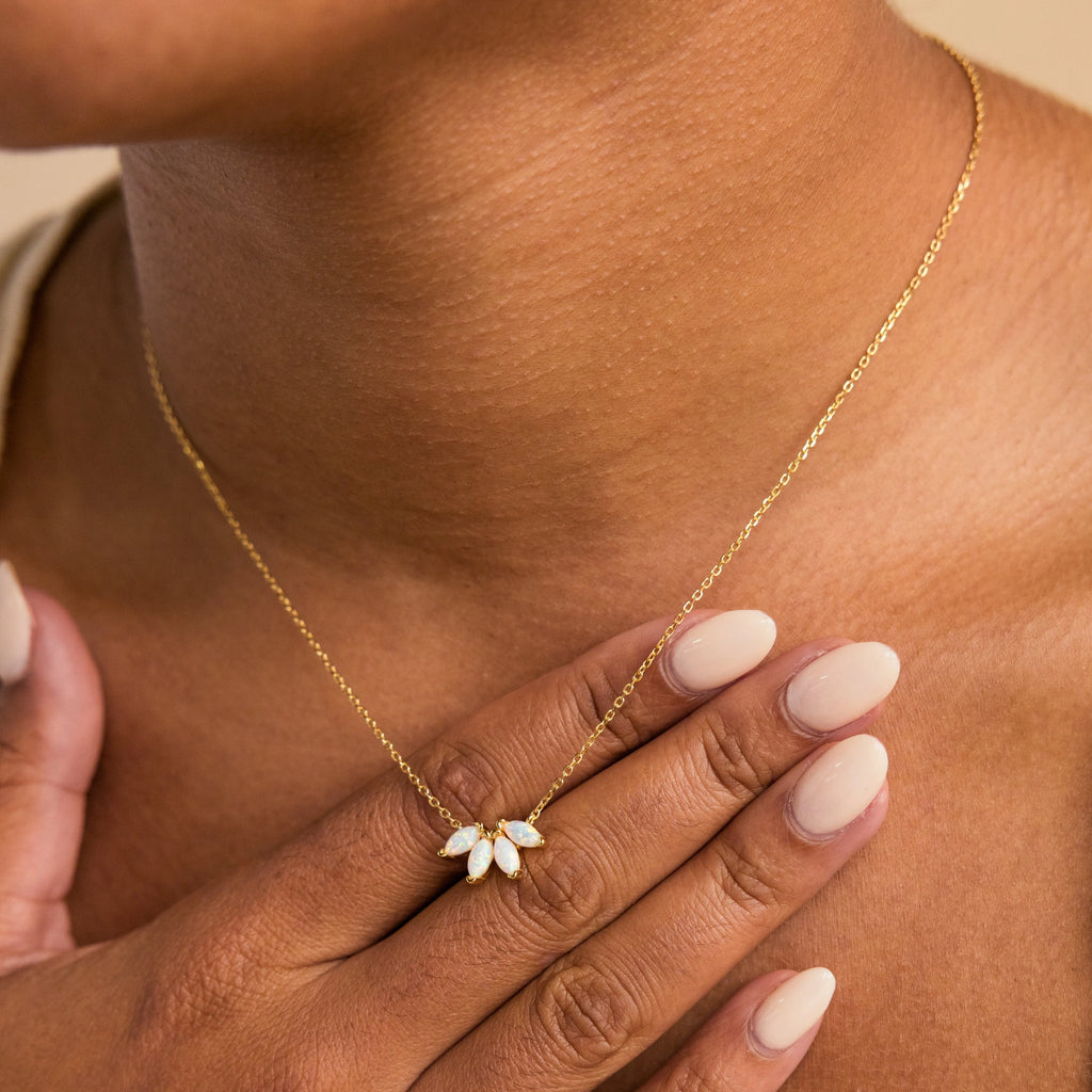 A person with manicured nails holds their chest, wearing the White Opal Marquise Necklace featuring a delicate, flower-shaped gold pendant.