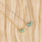 Two delicate necklaces rest on beige textured fabric: one features a gold floral design with a blue-green gemstone pendant, and the other is the Turquoise Opal Marquise Necklace in silver, both exuding understated elegance.