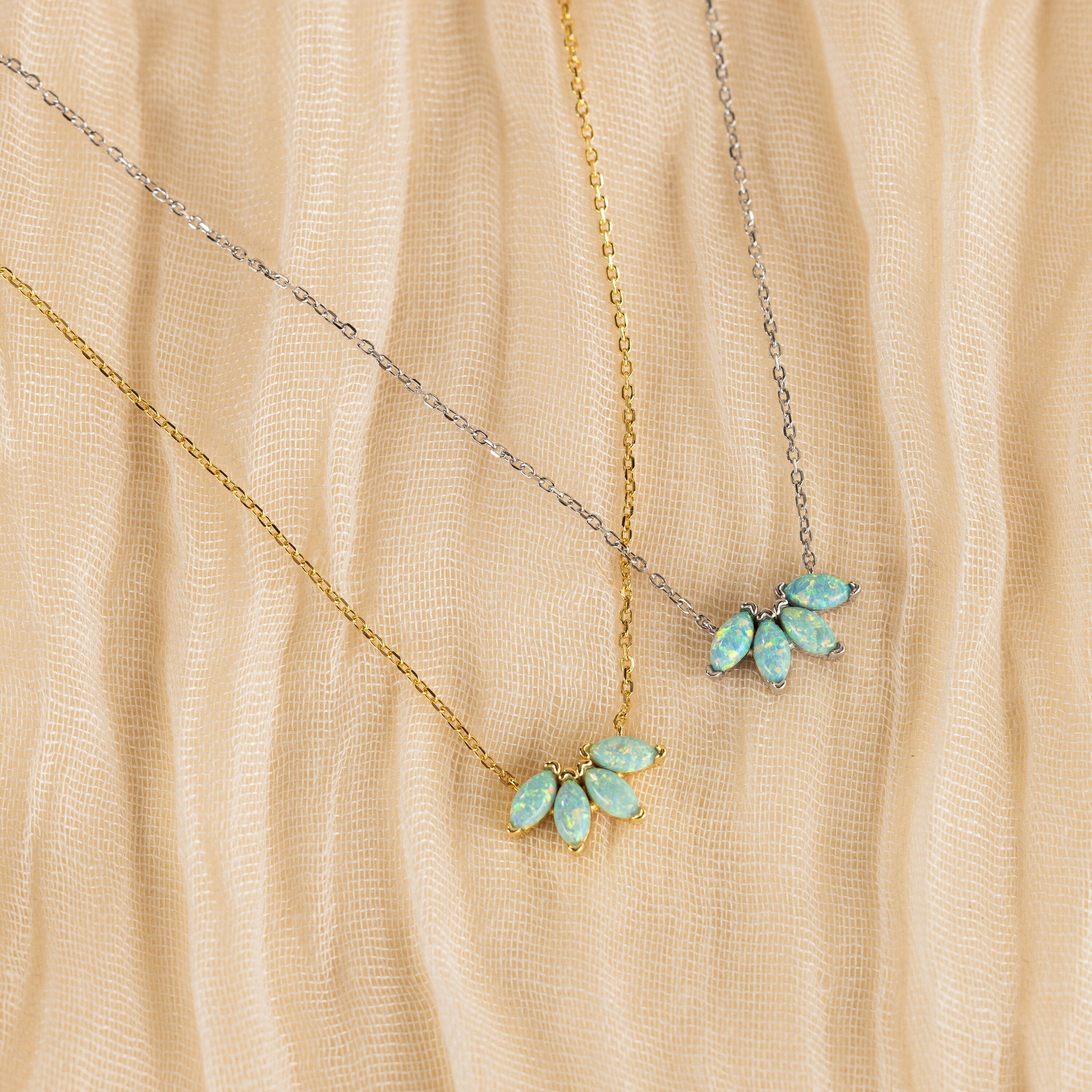 Two delicate necklaces rest on beige textured fabric: one features a gold floral design with a blue-green gemstone pendant, and the other is the Turquoise Opal Marquise Necklace in silver, both exuding understated elegance.