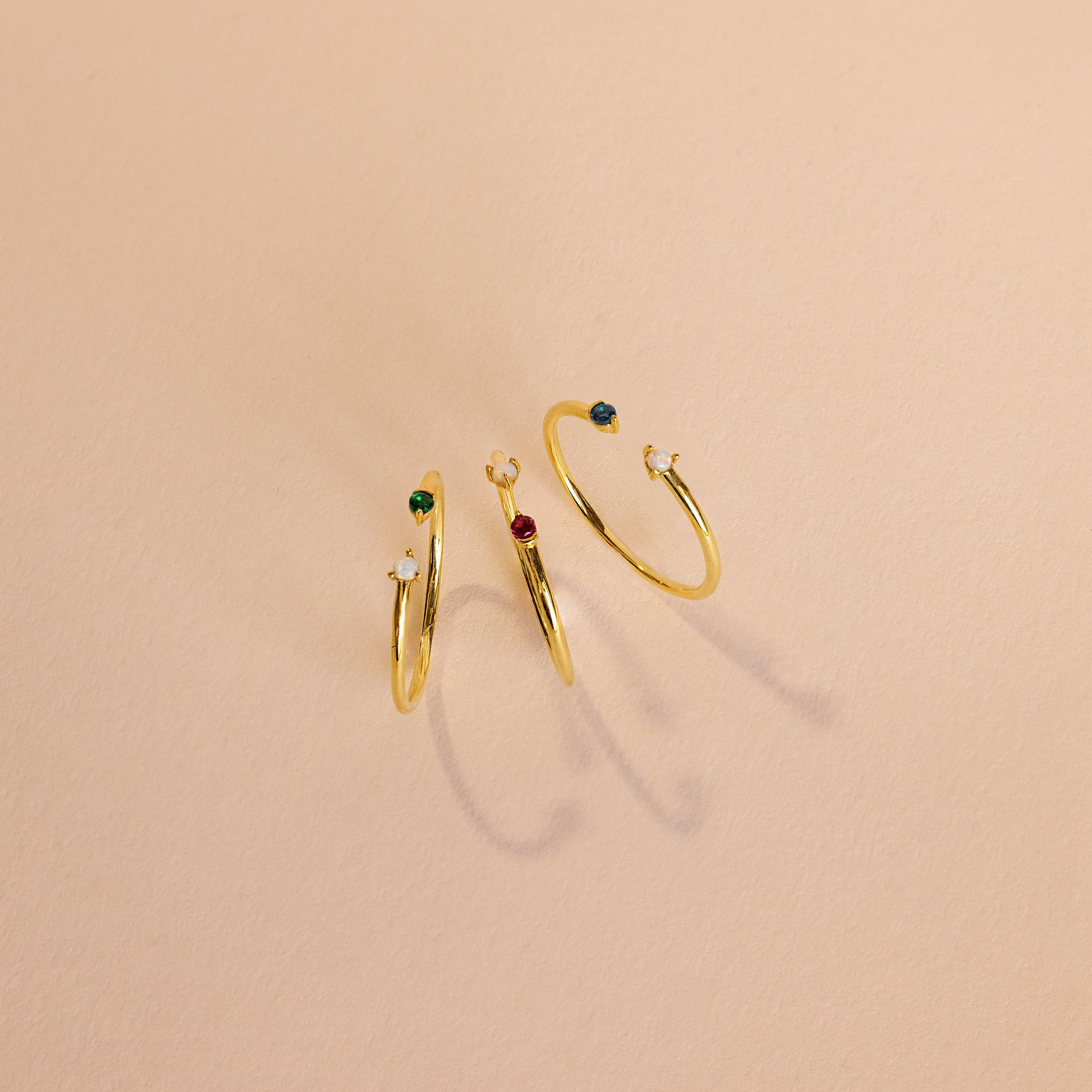 The Duo Birthstone & Opal Ring and two gold rings with small colored gemstones are displayed on a light beige surface, casting soft shadows.