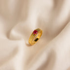 The Ribbed Birthstone Signet Ring features a ribbed gold band and a red gemstone, displayed on soft, cream-colored fabric.