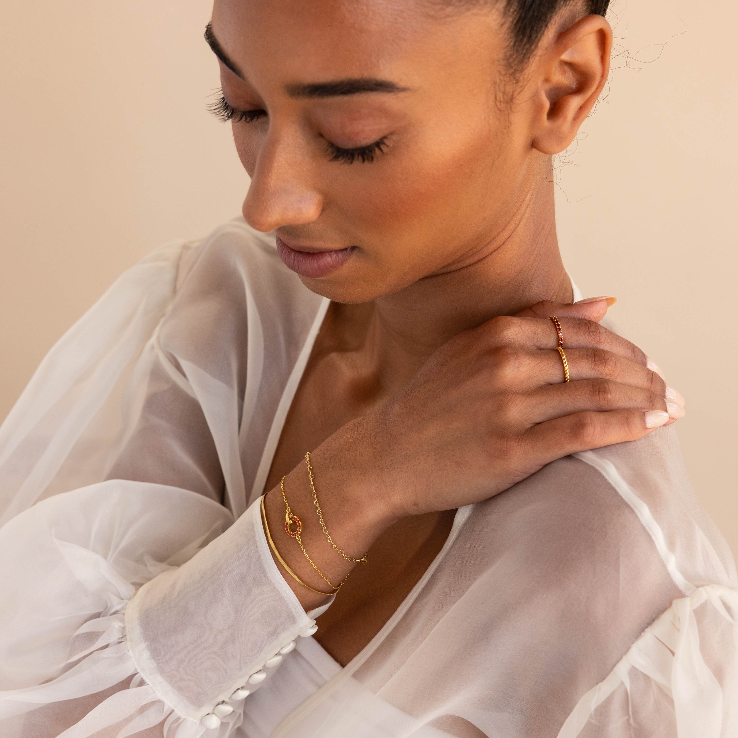 A woman in a sheer white blouse wears delicate gold rings and the Interlocking Birthstone Bracelet, gazing down with a gentle expression.