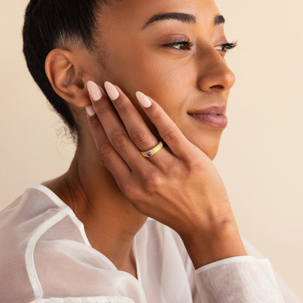 A woman with manicured nails smiles softly, resting her hand on her face and wearing the Ribbed Birthstone Signet Ring.