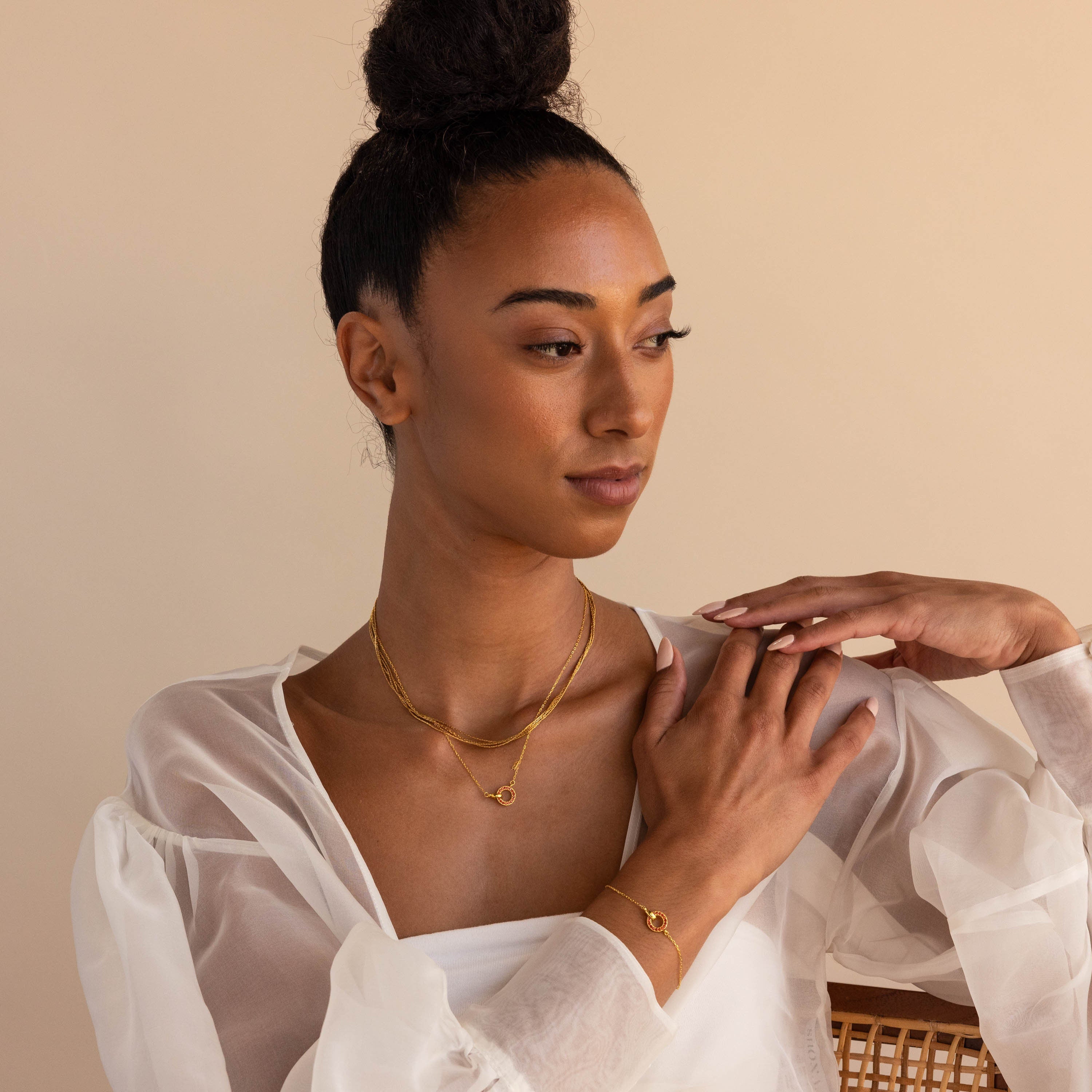 A woman wearing a sheer white blouse and the Interlocking Birthstone Necklace gazes thoughtfully to the side with her hand on her shoulder—a meaningful gift for mom.