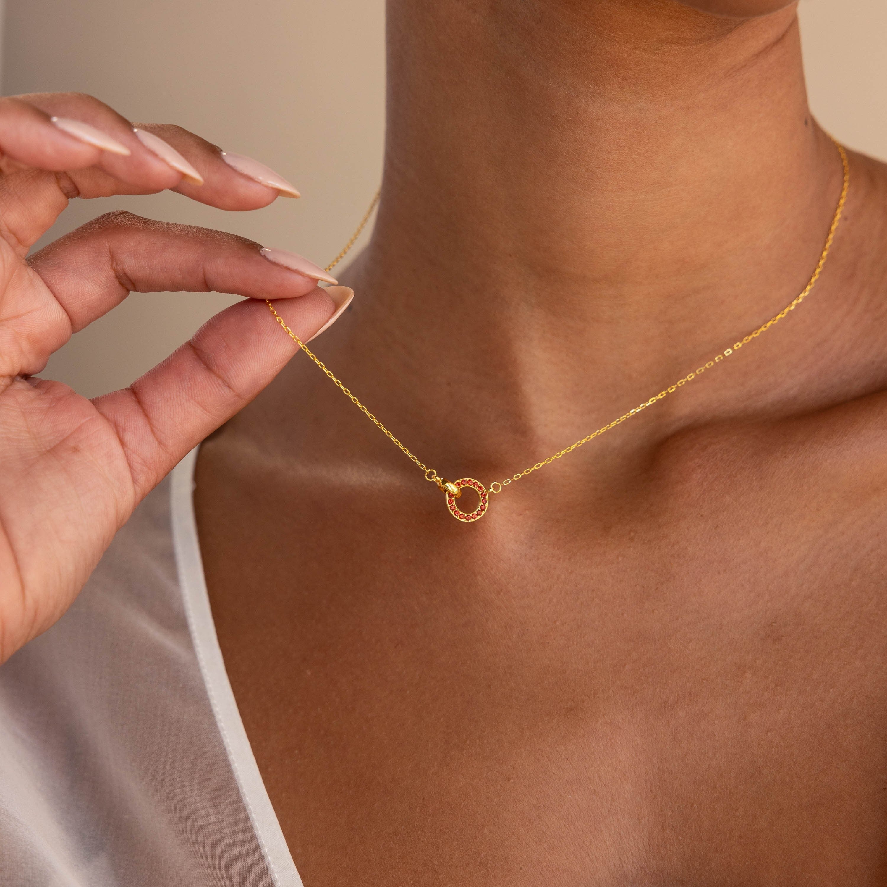 A woman wearing a white top holds the Interlocking Birthstone Necklace, featuring a delicate gold chain and small circular pendant—a perfect keepsake jewelry gift for mom.