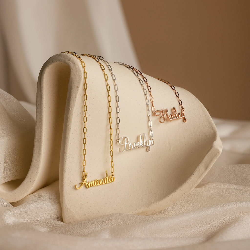The Luna Name Necklace in Paperclip Chain is shown with the names Amanda, Brooklyn, and Hazel on a curved off-white stand, each elegantly personalized on a chic paperclip chain.