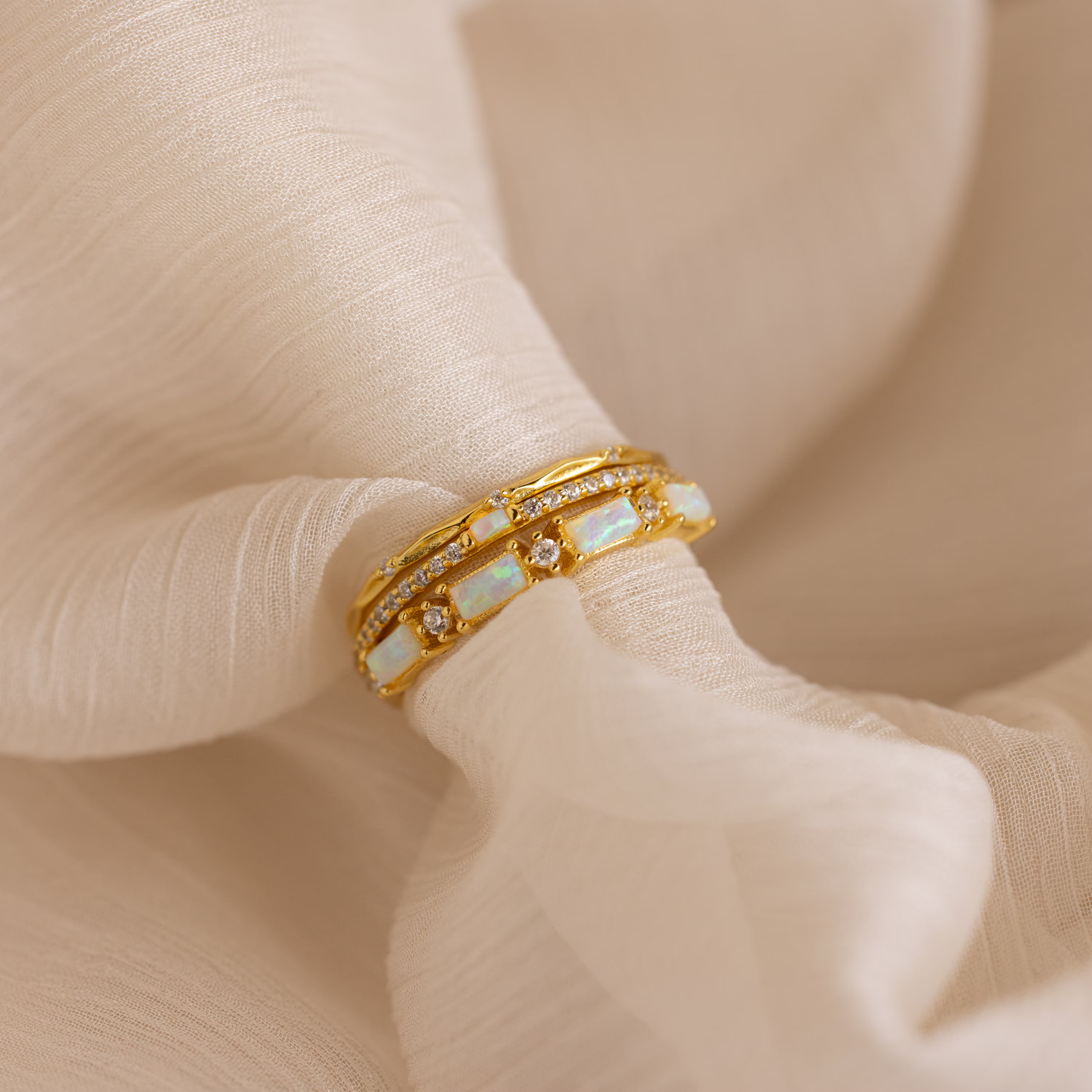 The Pave Opal Ring Set, featuring opal and diamond accents in a classic gold design, rests on soft, light beige fabric.