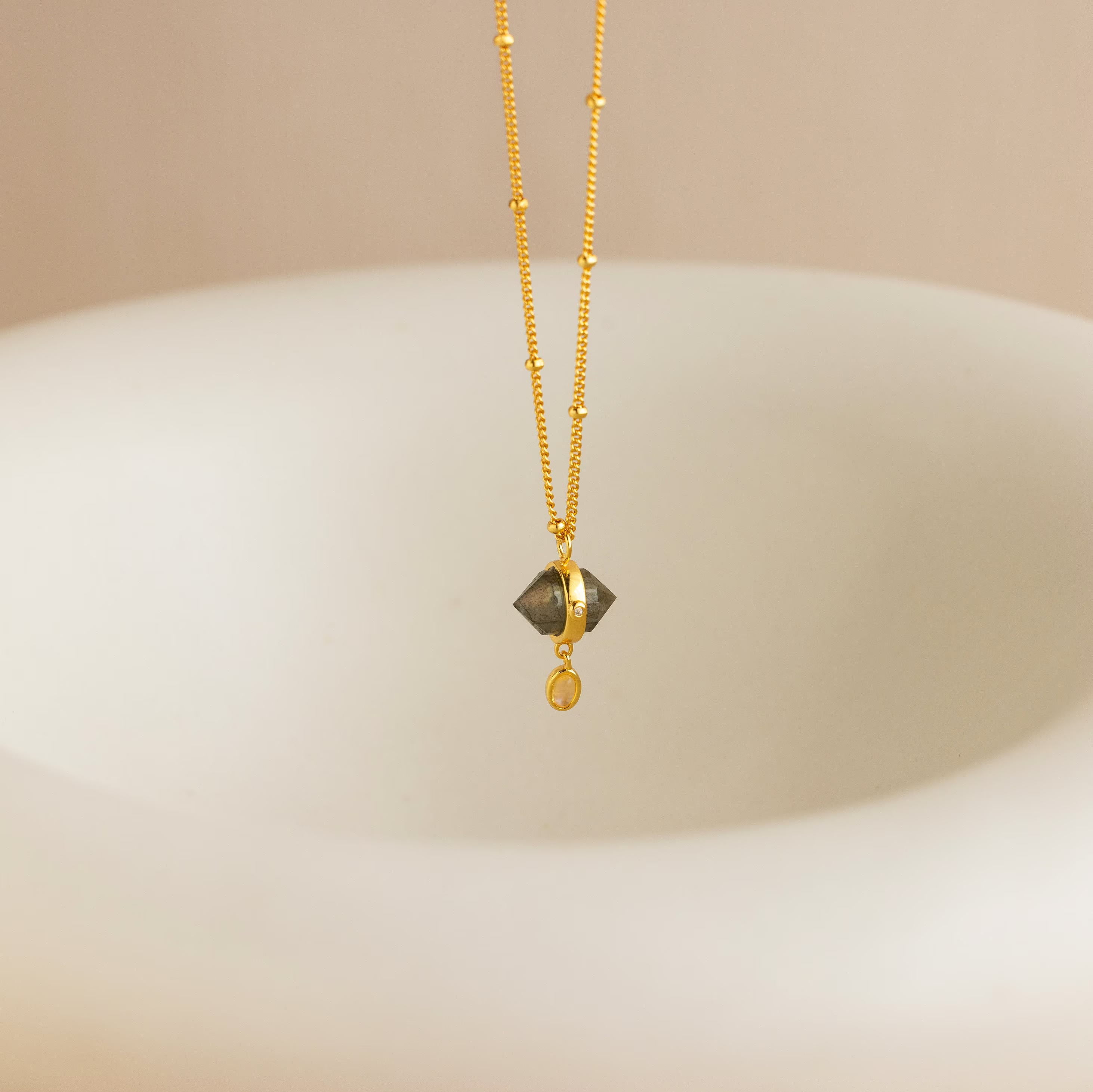 The Labradorite & Moonstone Drop Necklace features a shimmering gemstone pendant, suspended above a smooth, light-colored curved surface.