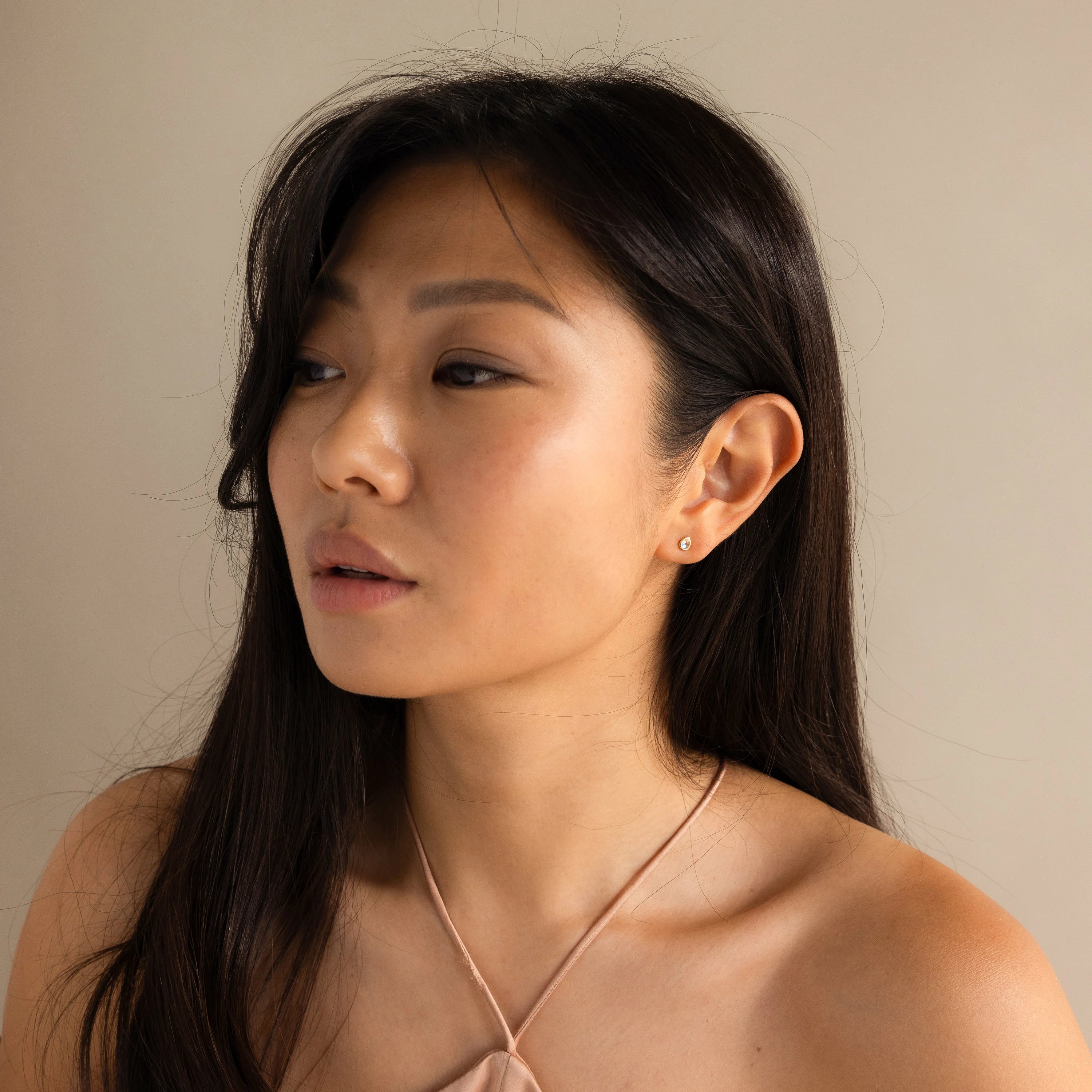 Woman with long dark hair and neutral expression looking to the side, wearing a light halter top and Pear Moonstone Studs that add a touch of celestial charm.