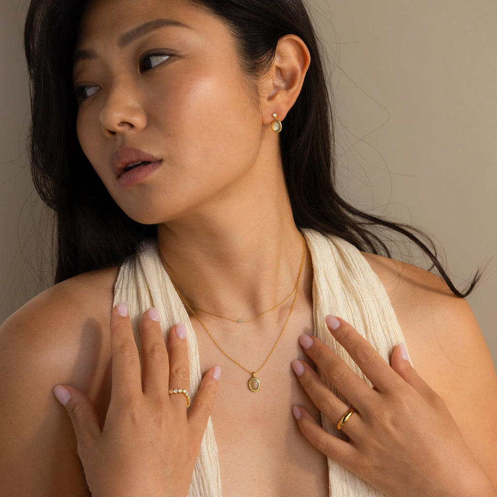 Woman with long dark hair wearing a cream sleeveless top and gold jewelry, including the Oval Labradorite Pendant Necklace, gazes to the side.