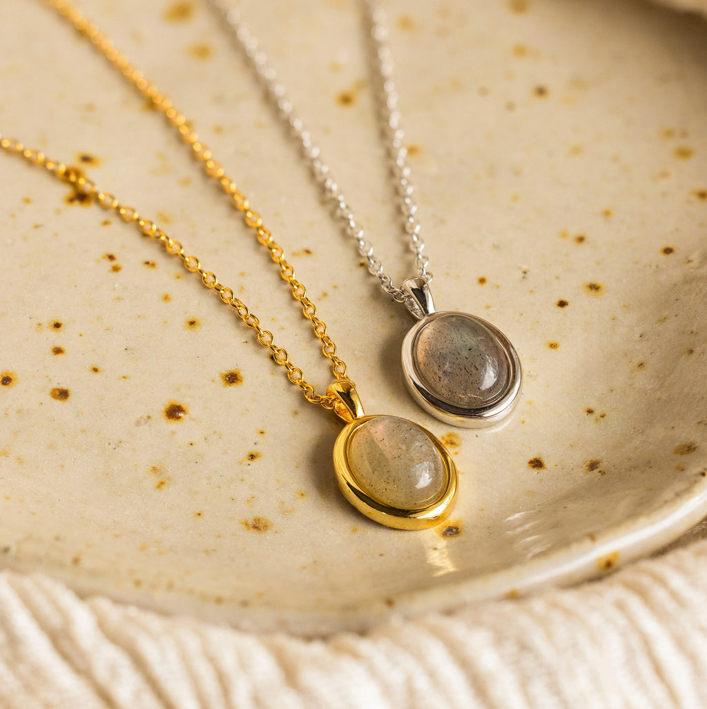 Two oval pendants, one gold and one silver, rest on a speckled ceramic surface. The Oval Labradorite Pendant Necklace highlights the unique shimmer and beauty of its labradorite gemstone.