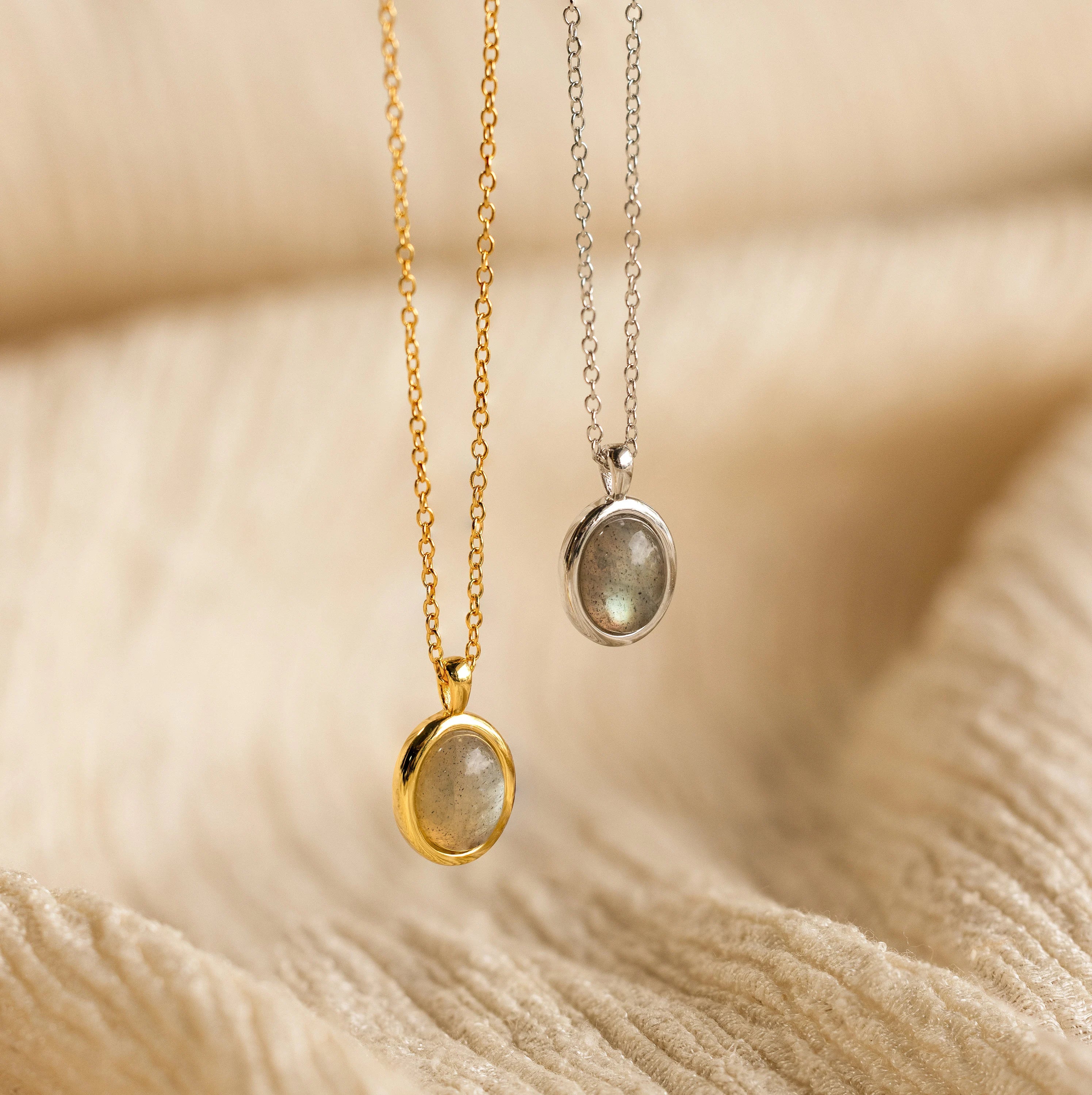 The Oval Labradorite Pendant Necklace features an oval pendant resting on a gold and silver chain, displayed against soft beige fabric.