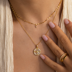 A woman showcases layered gold necklaces, featuring the Pave Pearl Compass Necklace, and a gemmed gold ring. Her manicured, nude-colored nails complete this elegant nautical-inspired jewelry look.