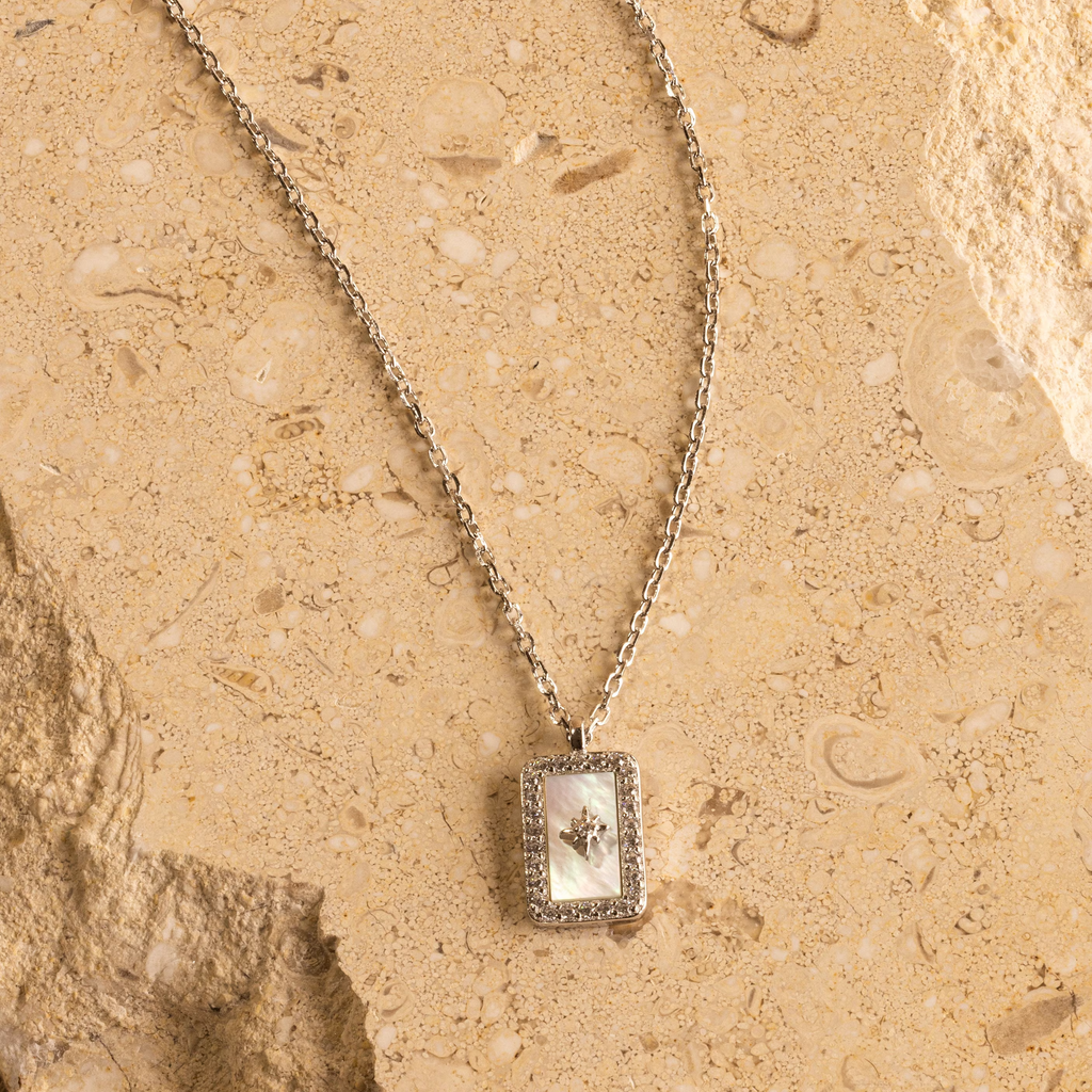The Pave Pearl North Star Necklace showcases a luminous center, set in silver and styled on a textured beige stone—an elegant celestial-inspired accessory.
