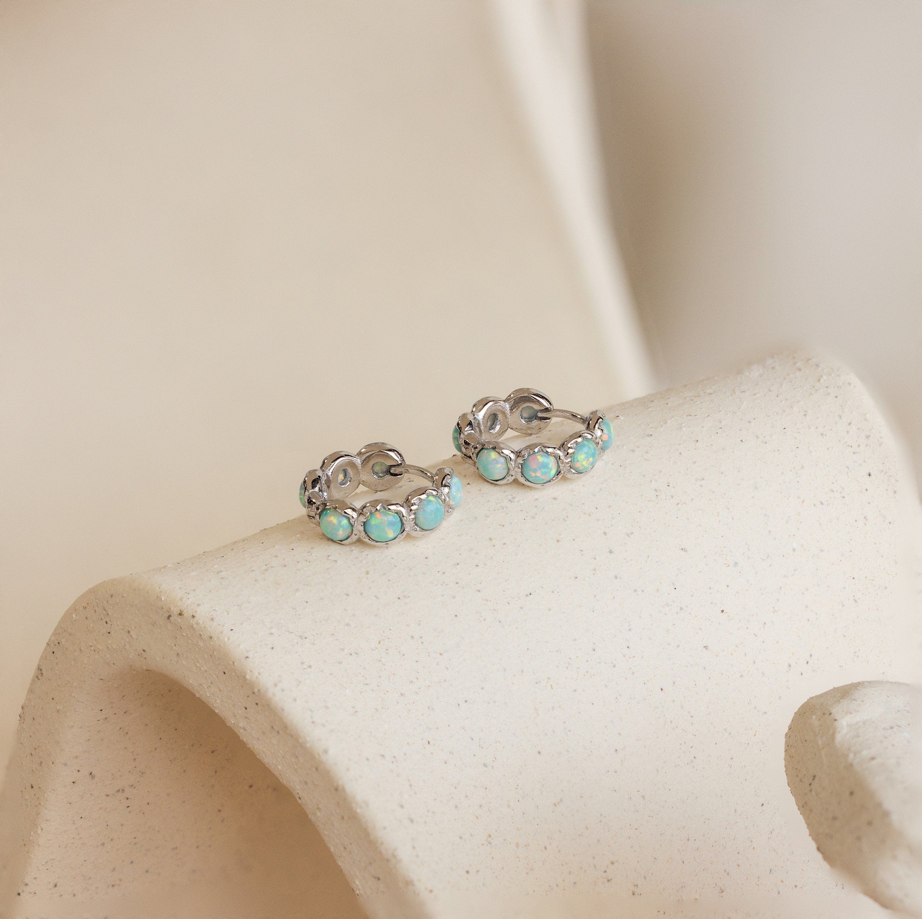 Opal Bezel Huggies featuring round blue opal stones, beautifully displayed on a textured, curved beige surface.