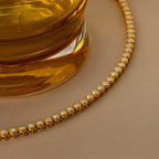 The Opal Bezel Tennis Necklace features round opal stones set in gold, beautifully capturing the elegance of a golden glass on a beige surface.