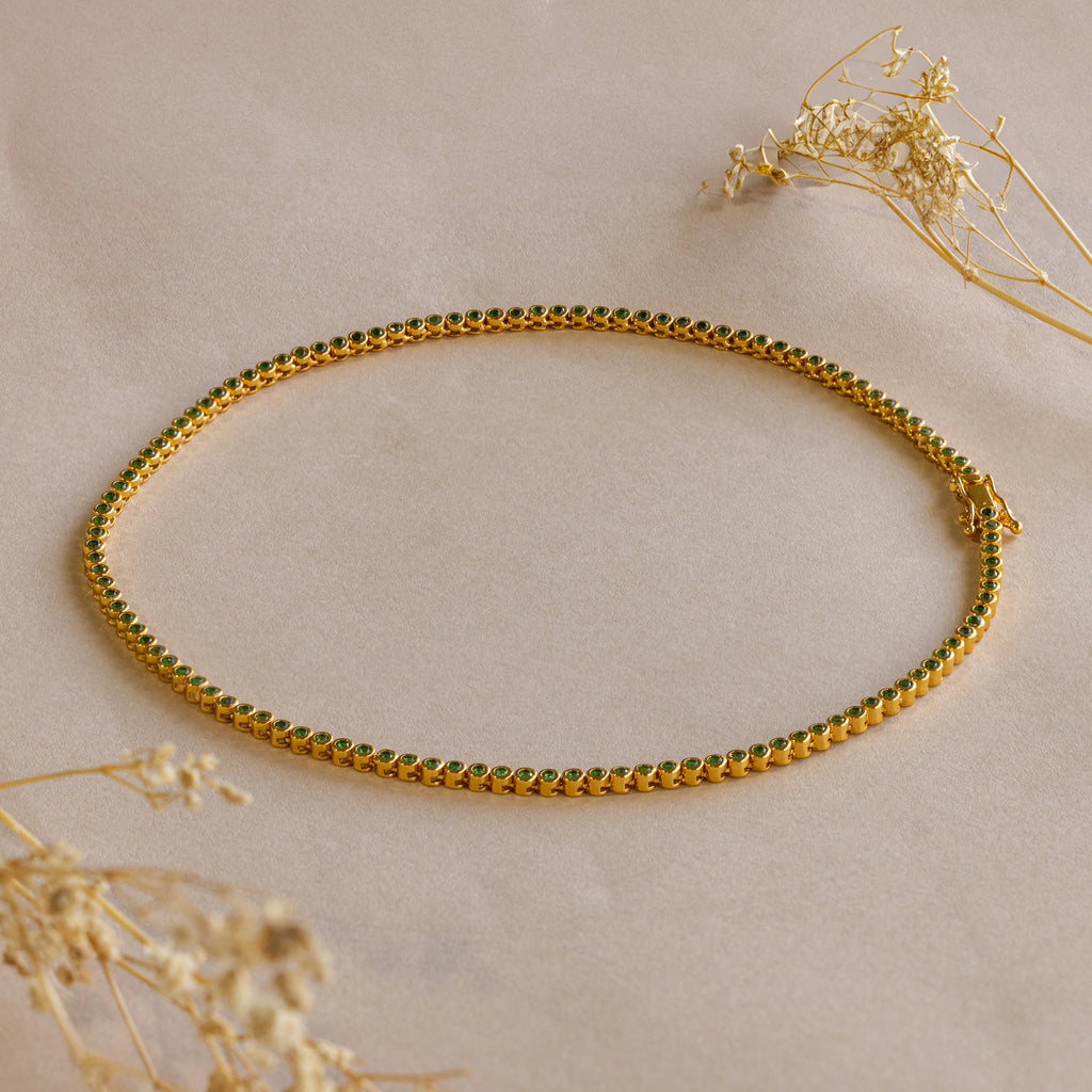 The Emerald Bezel Tennis Necklace, featuring gold and green accents, is displayed on a beige surface among dried flowers—a striking piece that brings elegance to any outfit.
