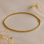 The Emerald Bezel Tennis Necklace, featuring gold and green accents, is displayed on a beige surface among dried flowers—a striking piece that brings elegance to any outfit.