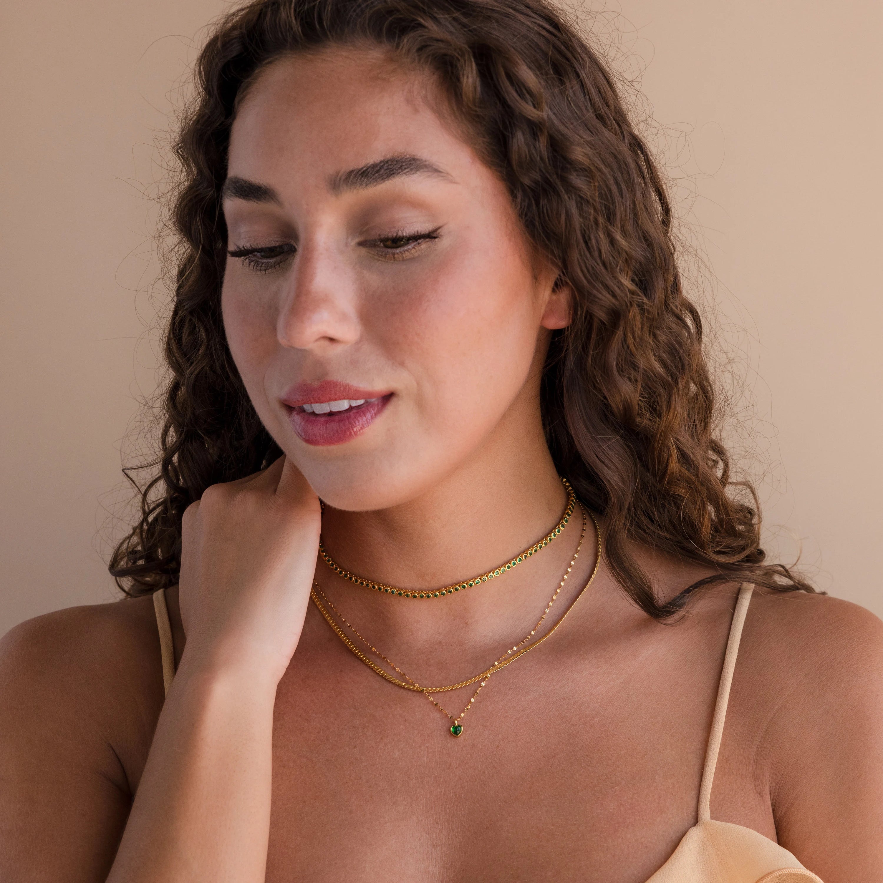 Woman with curly hair wears layered gold necklaces, featuring the elegant Emerald Bezel Tennis Necklace, paired with a beige spaghetti strap top as she looks down and smiles.