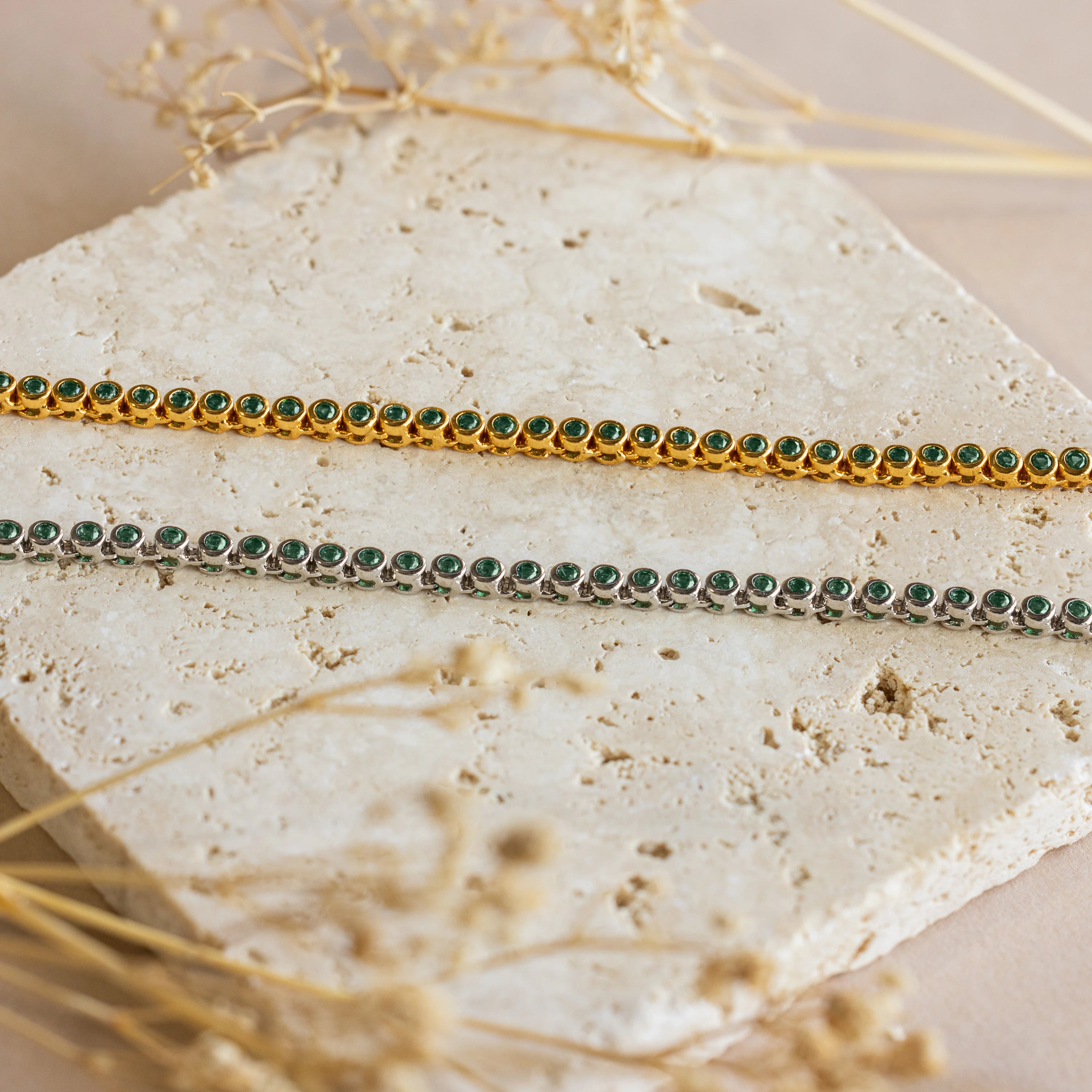 Two delicate bracelets—one gold, one silver with green gemstones—rest on stone beside the Emerald Bezel Tennis Necklace, ideal for pairing with your favorite cherished jewelry.