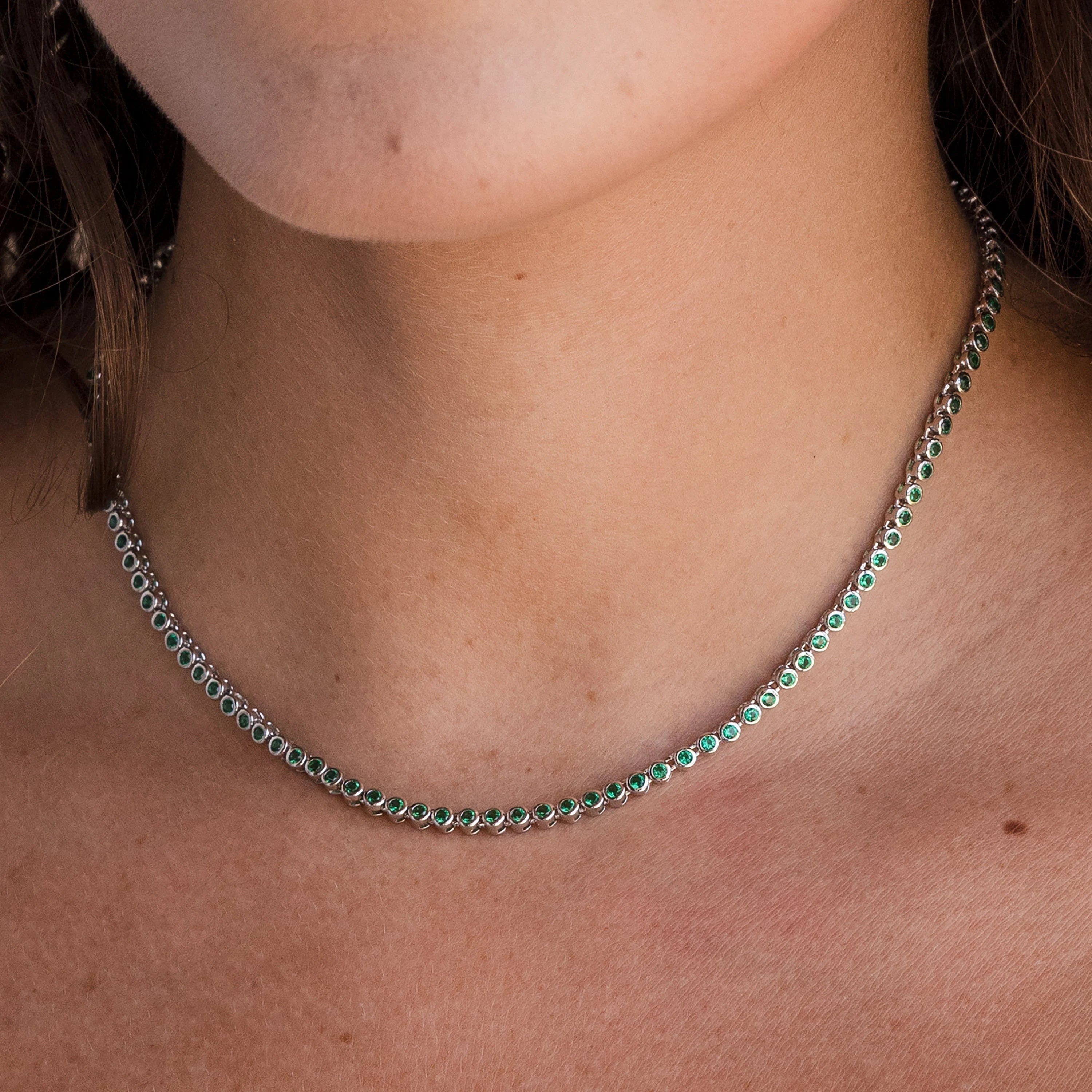 A woman wears the Emerald Bezel Tennis Necklace, showcasing petite green emerald stones set in silver, around her neck.