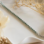 Emerald Bezel Tennis Necklace displayed on a wavy white surface with dried flowers—an elegant piece featuring green gemstones, perfect for any jewelry lover.