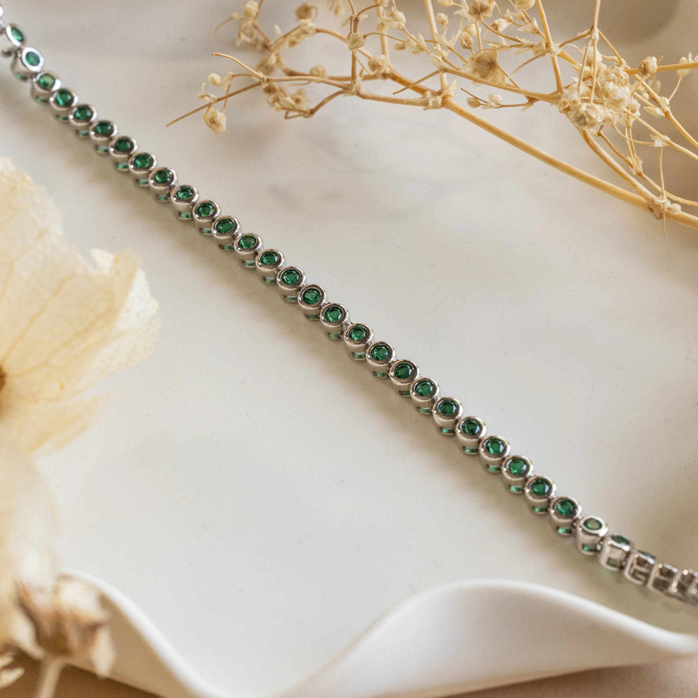 Emerald Bezel Tennis Necklace displayed on a wavy white surface with dried flowers—an elegant piece featuring green gemstones, perfect for any jewelry lover.