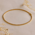 The Pink Bezel Tennis Necklace, adorned with pink gemstones, is displayed in a circle on a beige surface and accented with dried flowers.