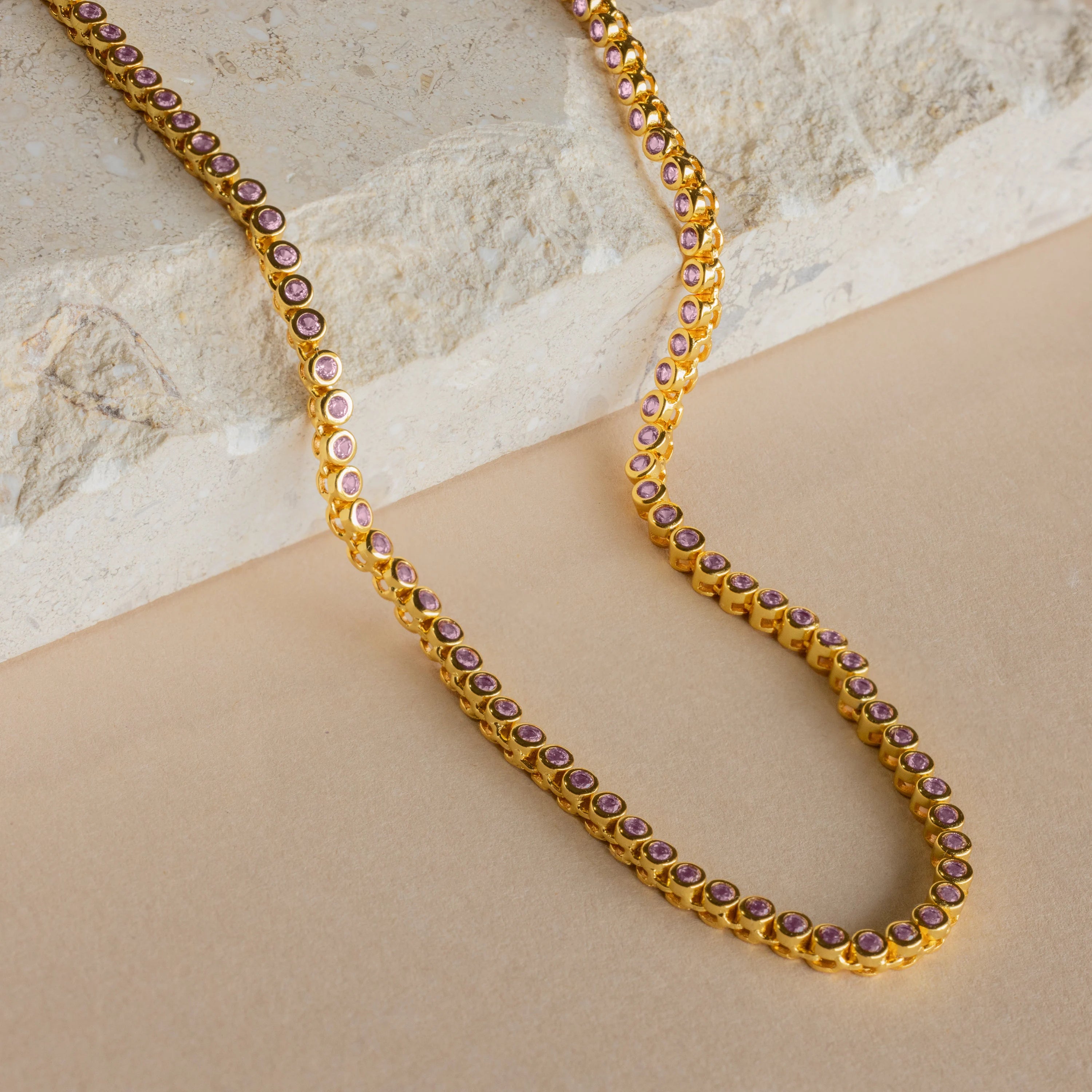 The Pink Bezel Tennis Necklace features elegant pink gemstones set in gold, artfully displayed on a beige stone and tan surface.