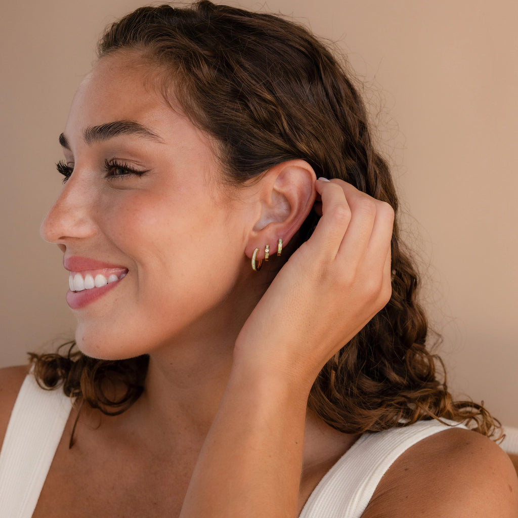 A smiling woman with curly hair touches her ear, showcasing the elegant Opal Inlay Diamond Huggies while wearing a white top.
