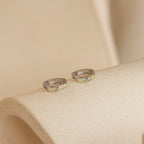 The Opal Inlay Diamond Huggies are elegant small silver hoop earrings featuring a single clear gemstone, beautifully displayed on a textured beige surface.