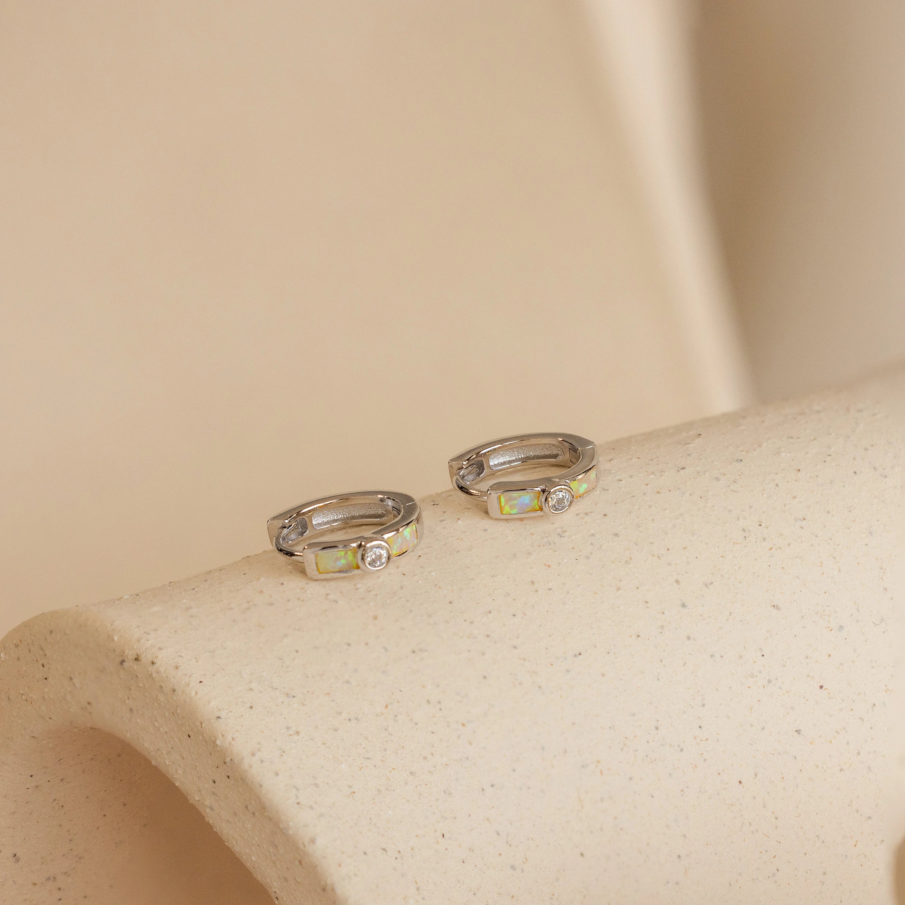 The Opal Inlay Diamond Huggies are elegant small silver hoop earrings featuring a single clear gemstone, beautifully displayed on a textured beige surface.