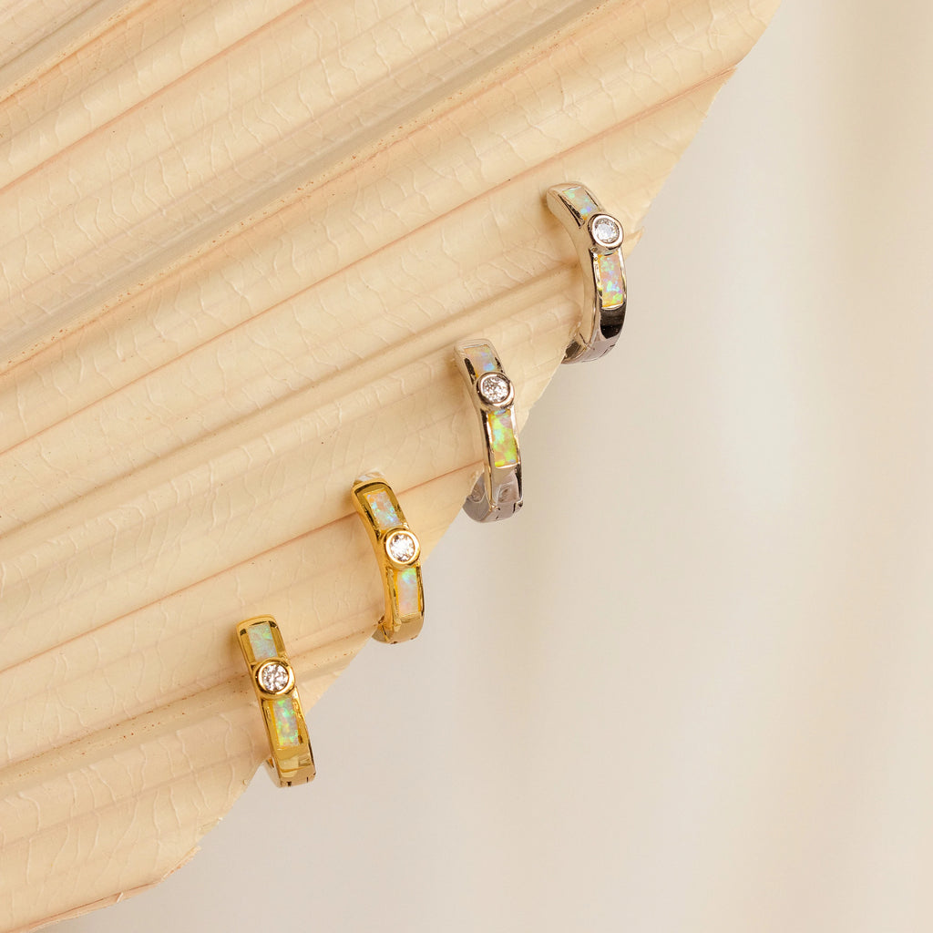 Three elegant Opal Inlay Diamond Huggies, featuring gold hoops with opal inlay and a small diamond accent, displayed on a textured beige surface.
